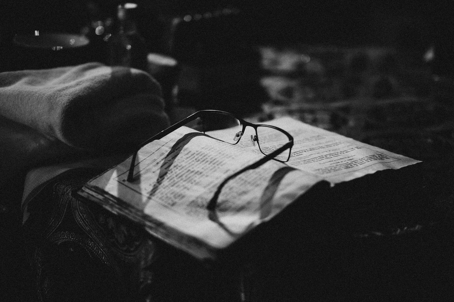 Black and white photo of eyeglasses resting on an open book in dim light.
