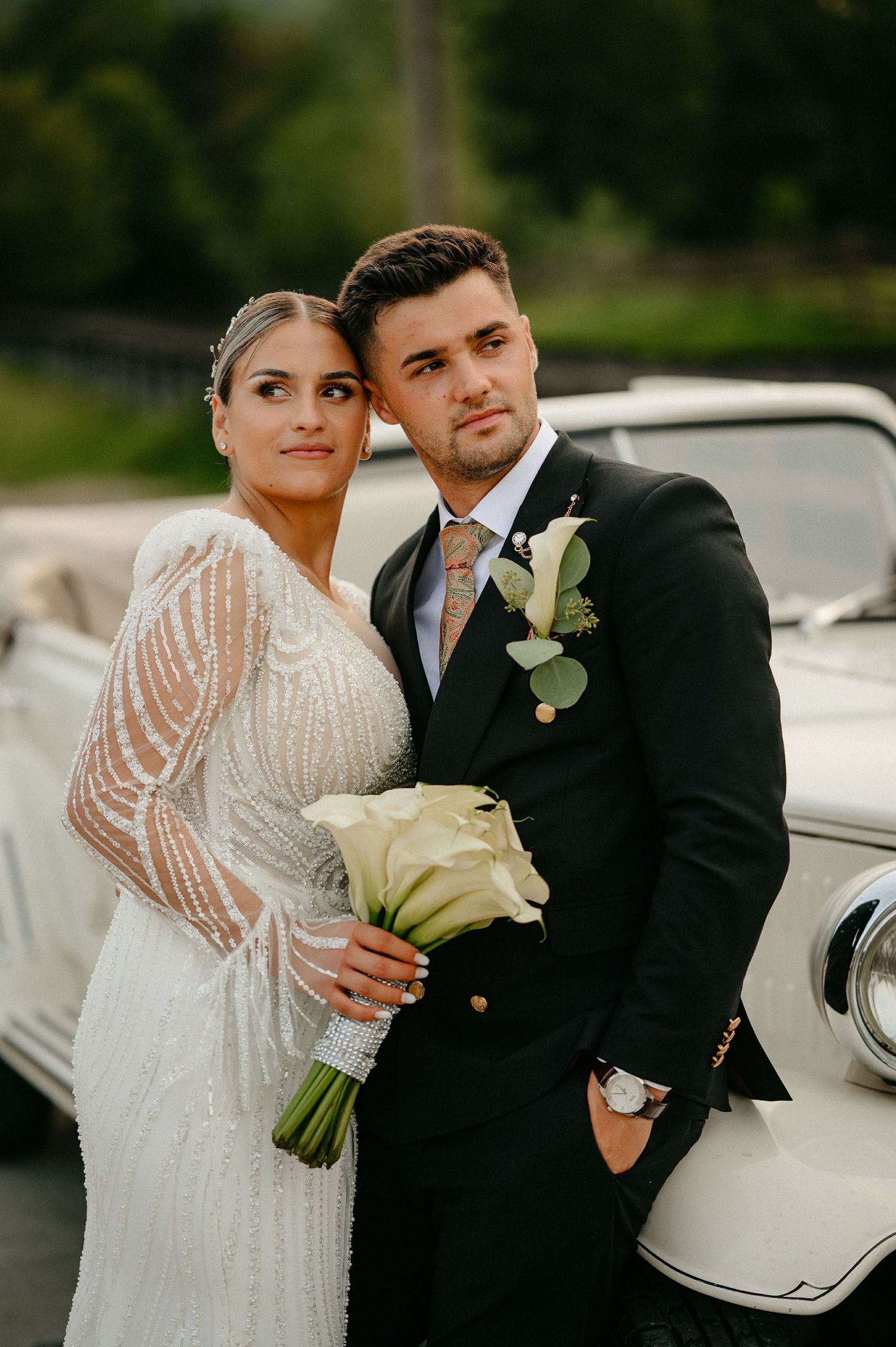 Stylish bride in beaded gown and groom in black suit leaning against a white vintage car.