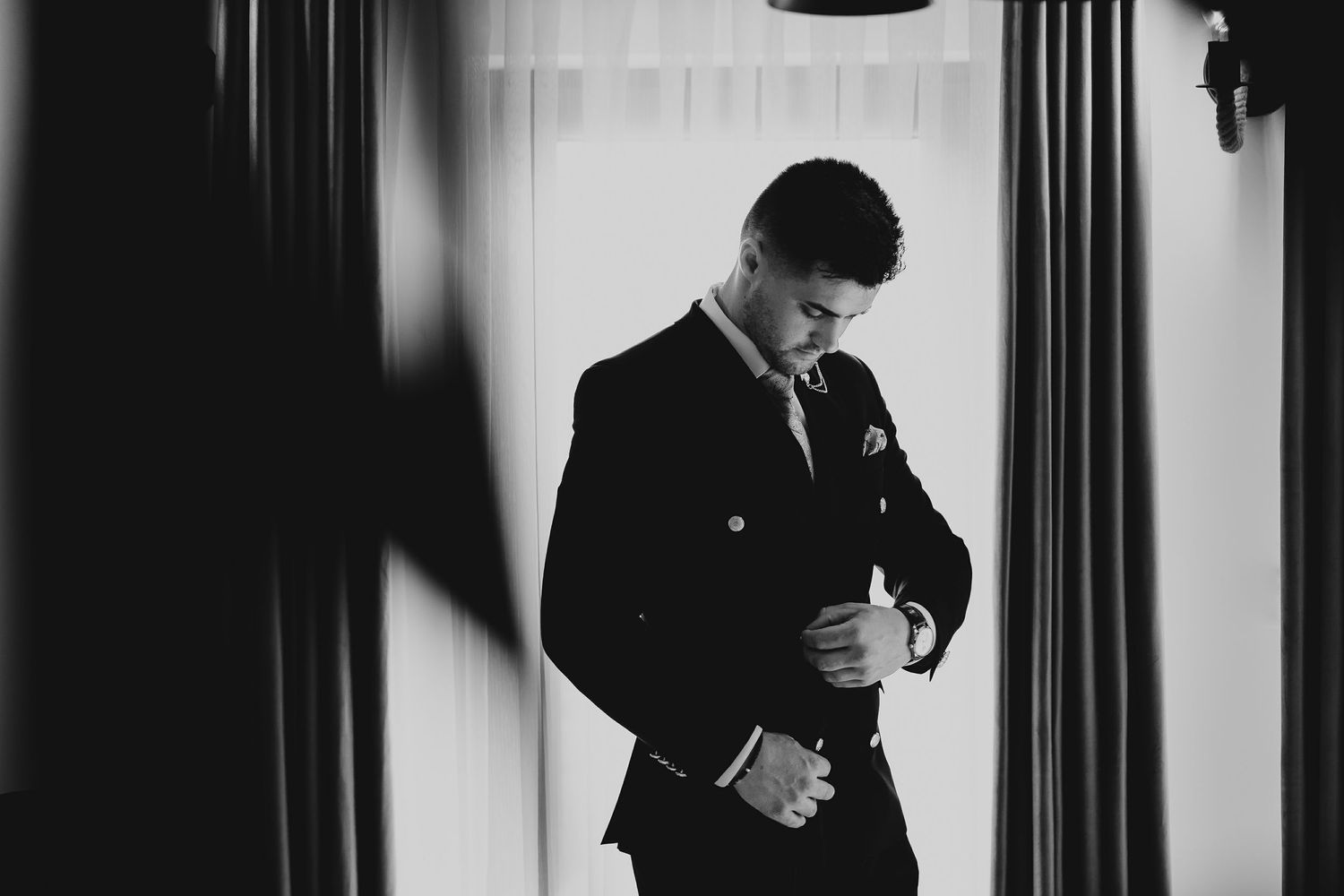 Black and white photo of groom in dark suit buttoning jacket by window.