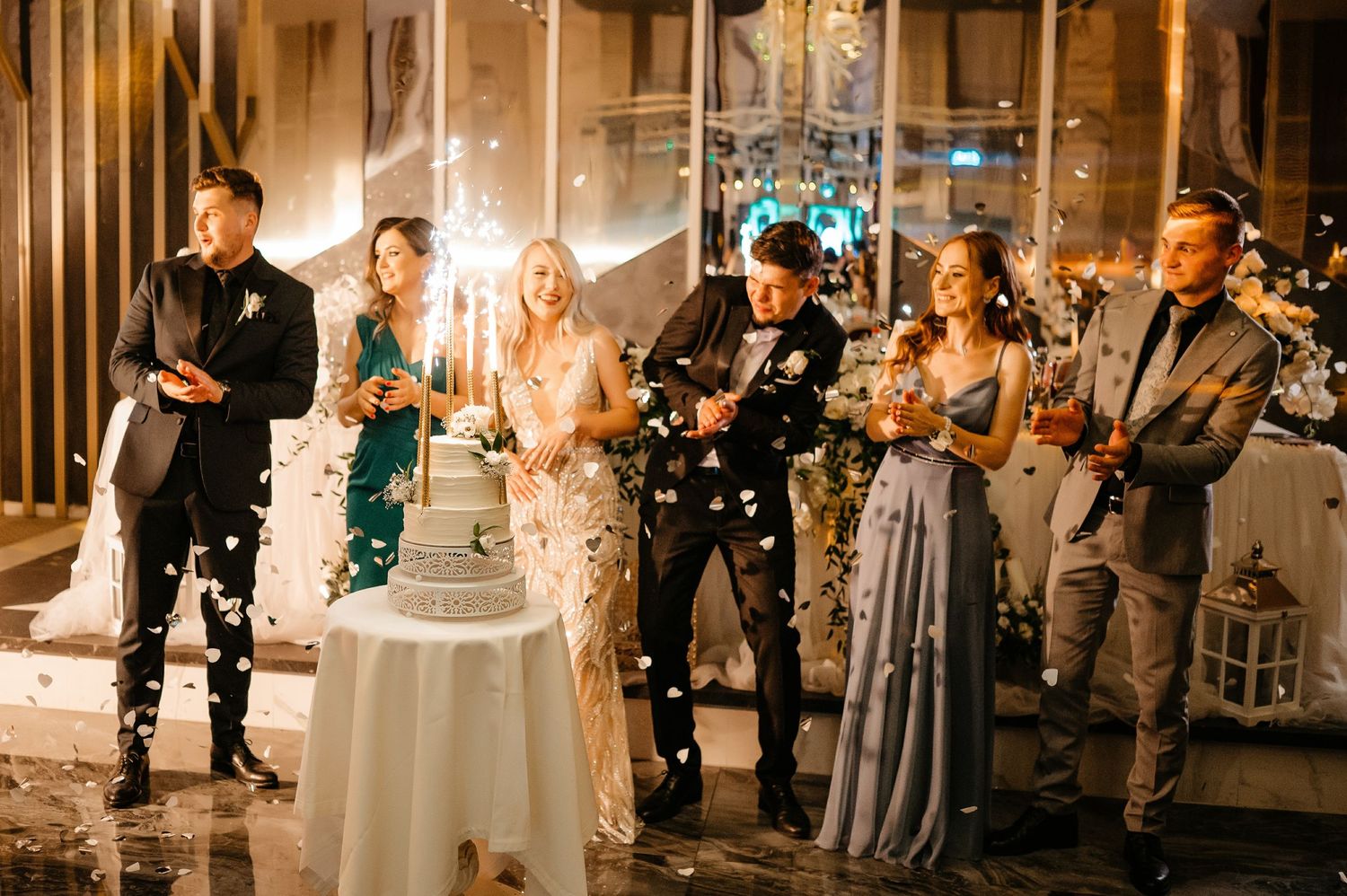Bride and groom cut wedding cake surrounded by guests with sparklers at evening reception.
