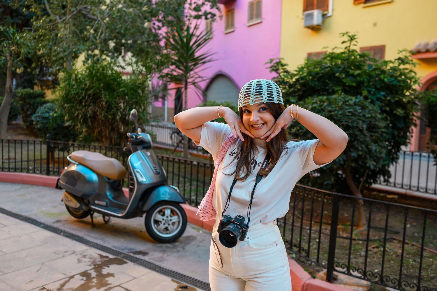 Smiling young woman wearing a colorful helmet and holding a camera, posing near a scooter outdoors.
