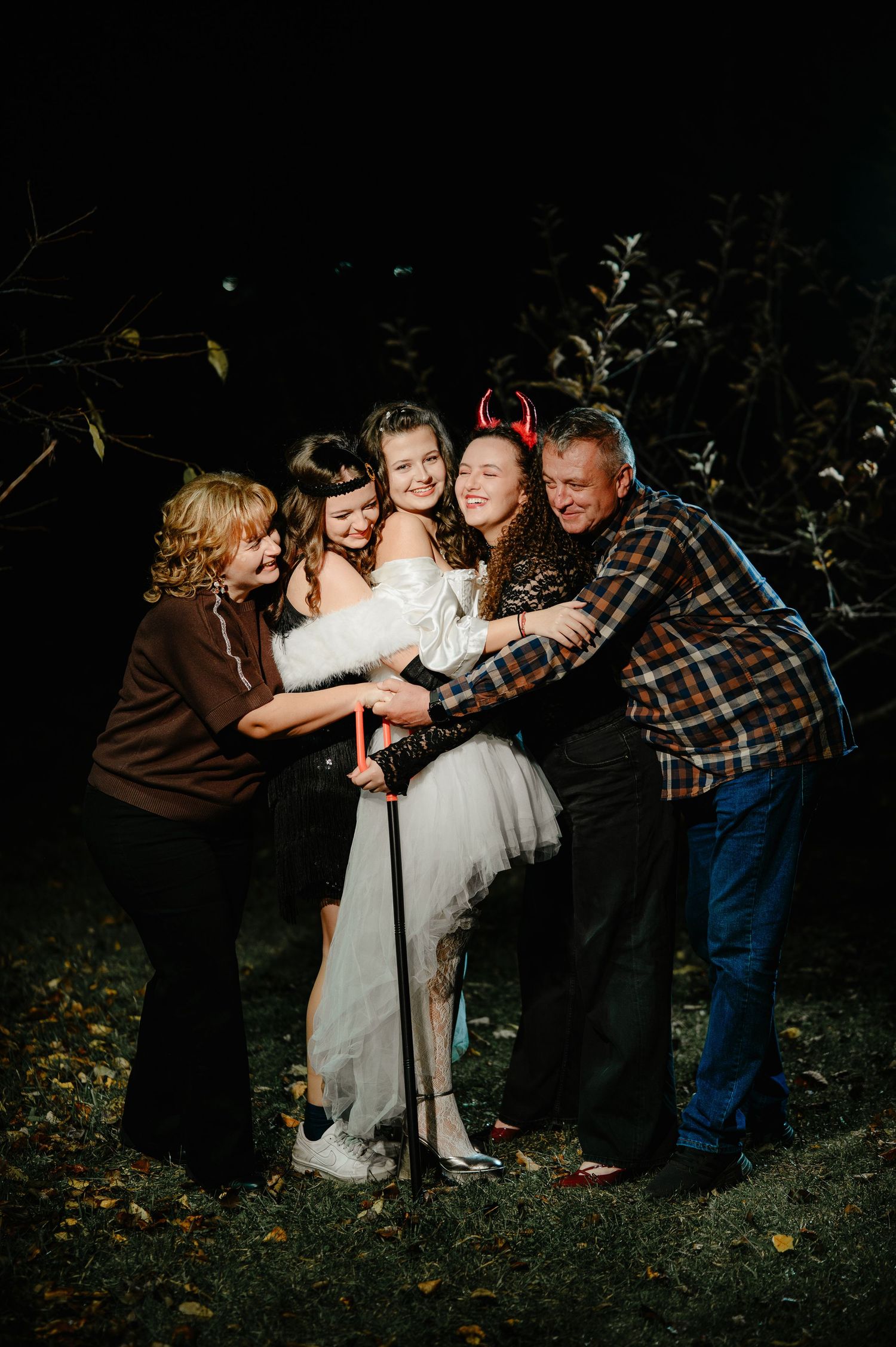 Group of five people hugging and celebrating together outdoors at night in Halloween costumes.
