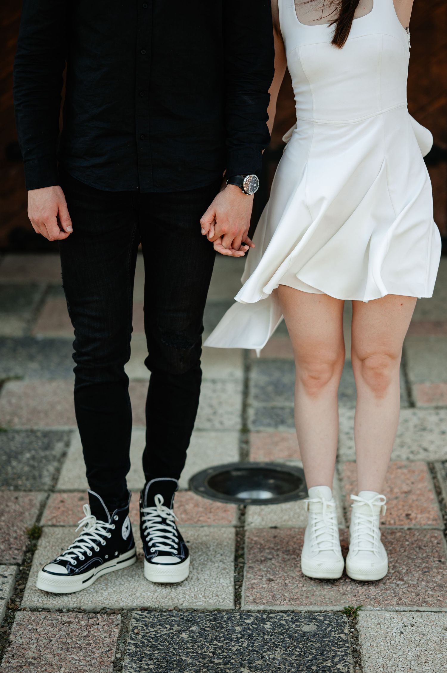 Close-up of couple holding hands, both wearing Converse sneakers on a cobblestone pavement.