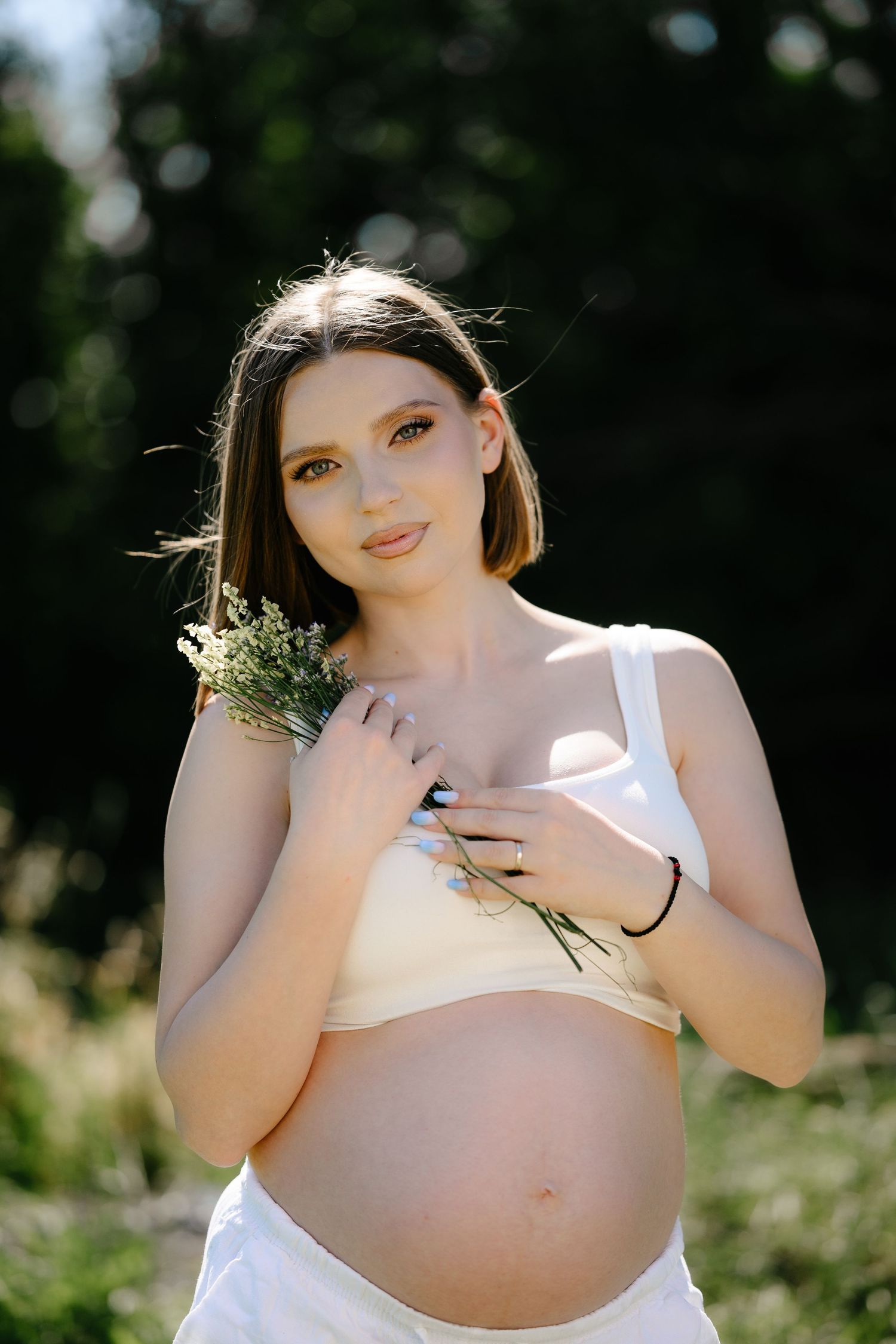 Smiling pregnant woman in white crop top holding wildflowers against her bare bump in sunny outdoor setting.