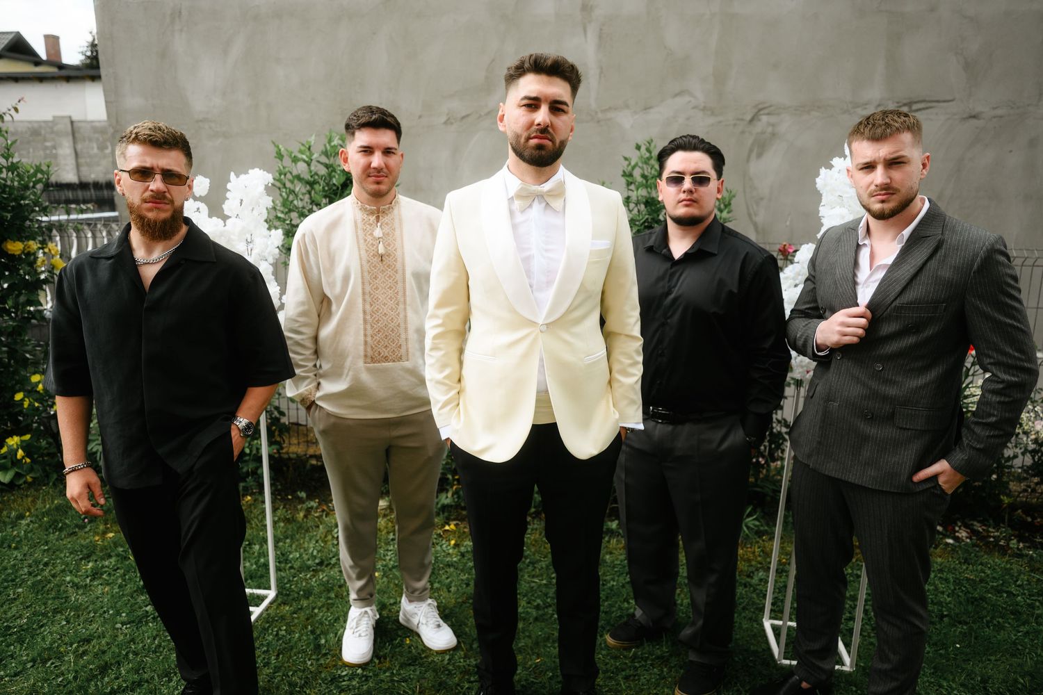 Five young men in formal attire pose outdoors, with the center man wearing a white suit at what appears to be a wedding.