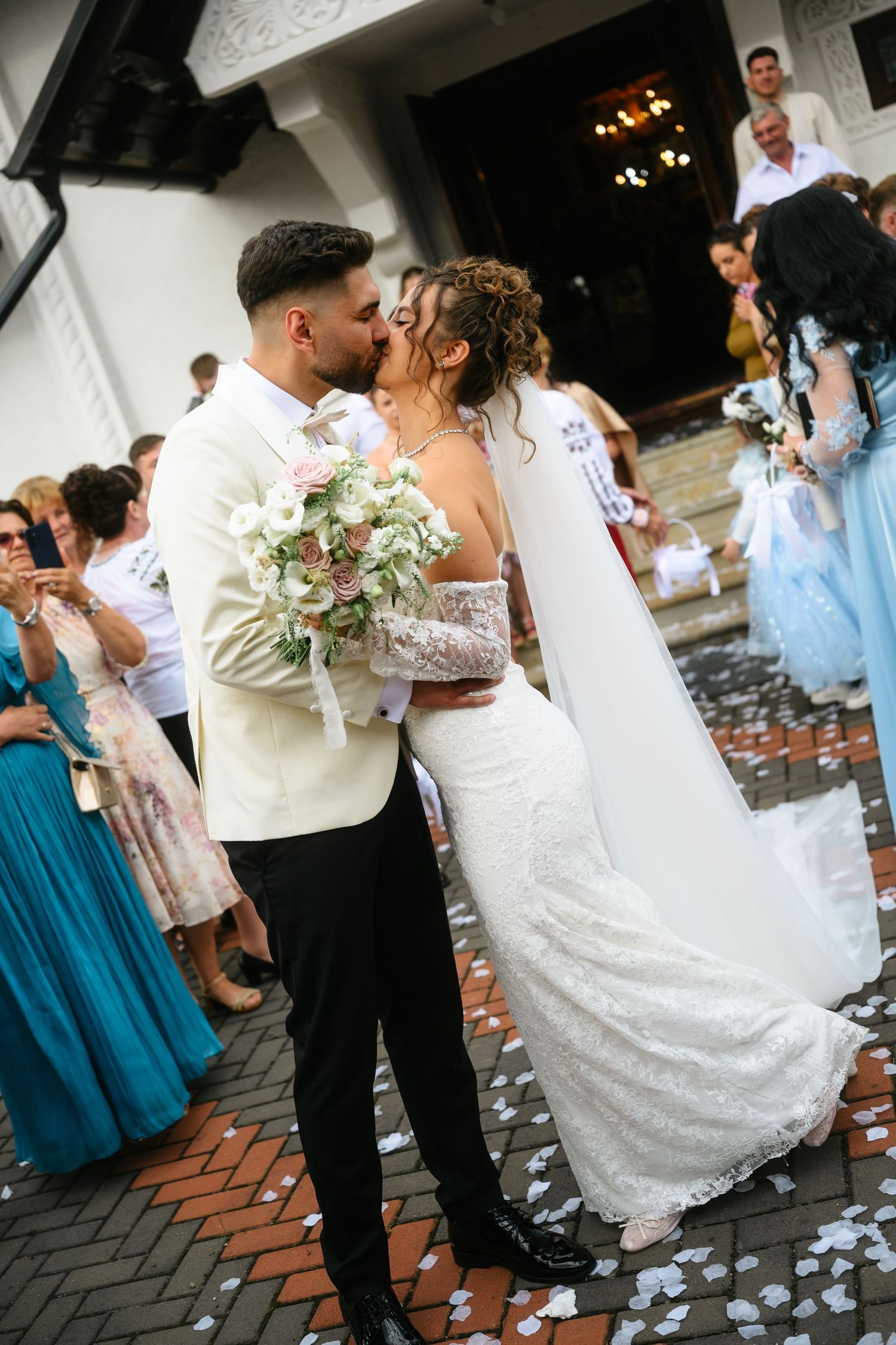 Bride and groom share a kiss outside church, surrounded by guests throwing white petals, bride holding pink bouquet.