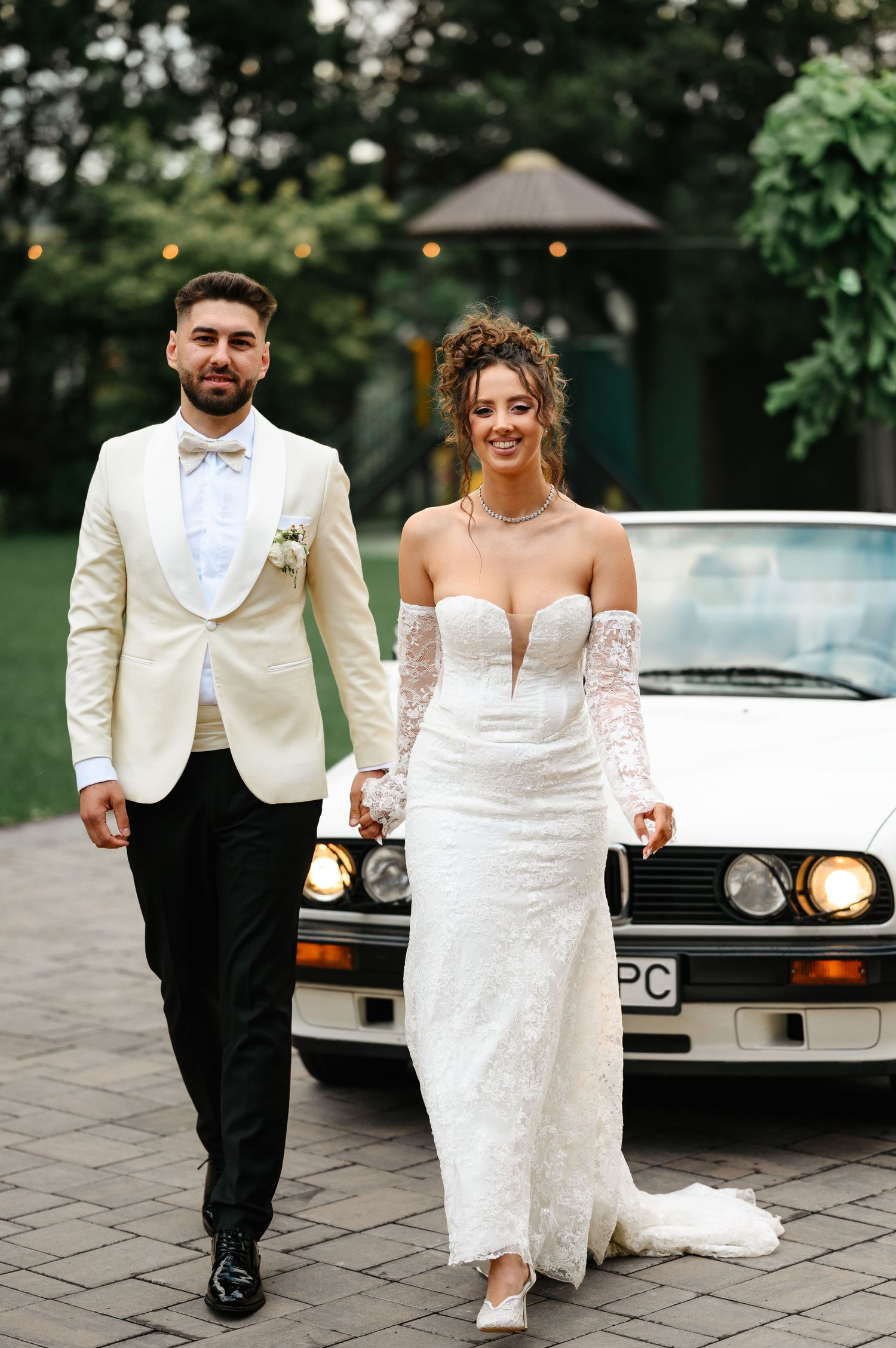 Bride and groom walking hand in hand in front of a white vintage car, wearing elegant white wedding attire.