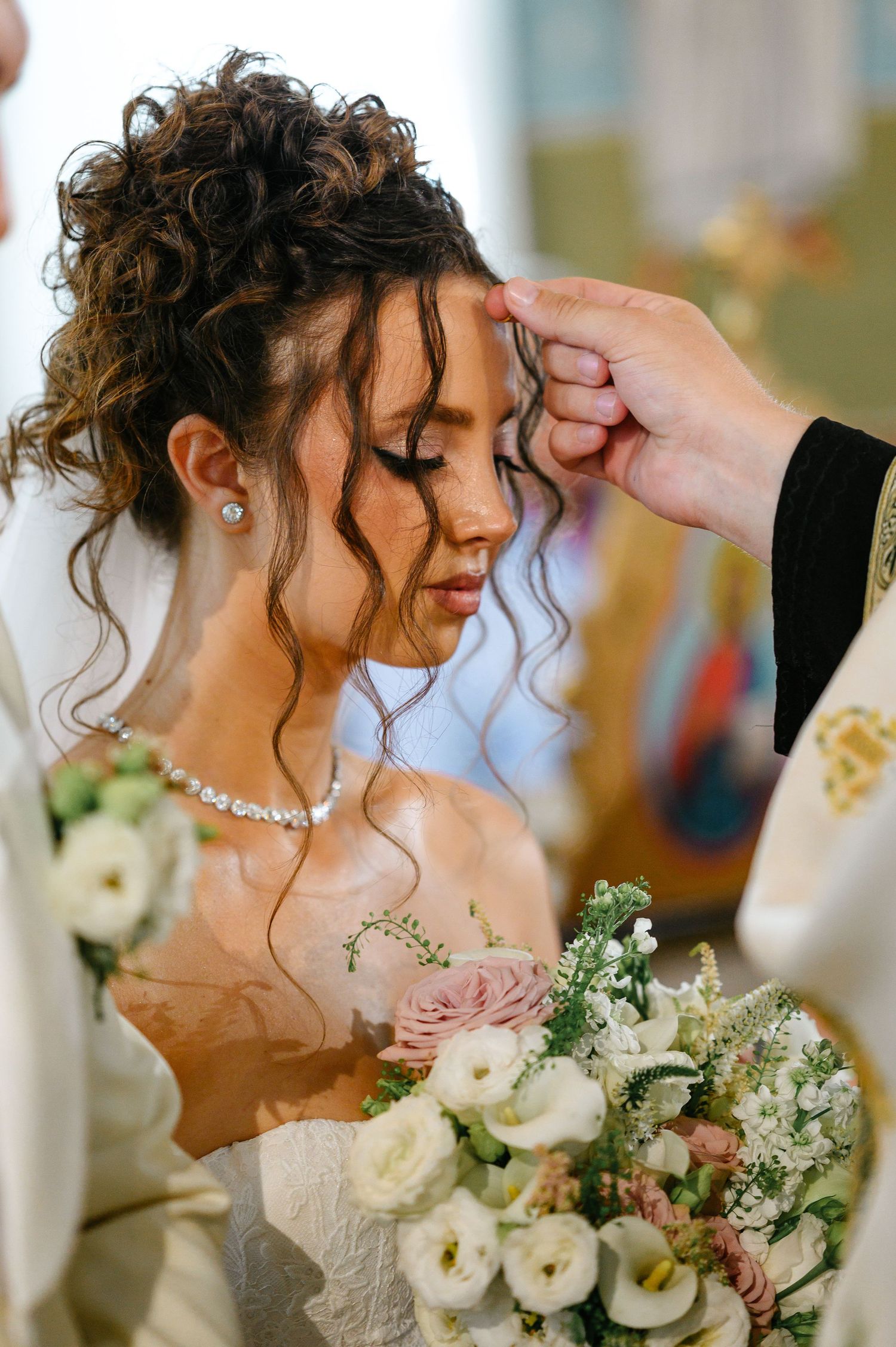 Bride in white dress holds floral bouquet during church wedding ceremony while officiant blesses her.