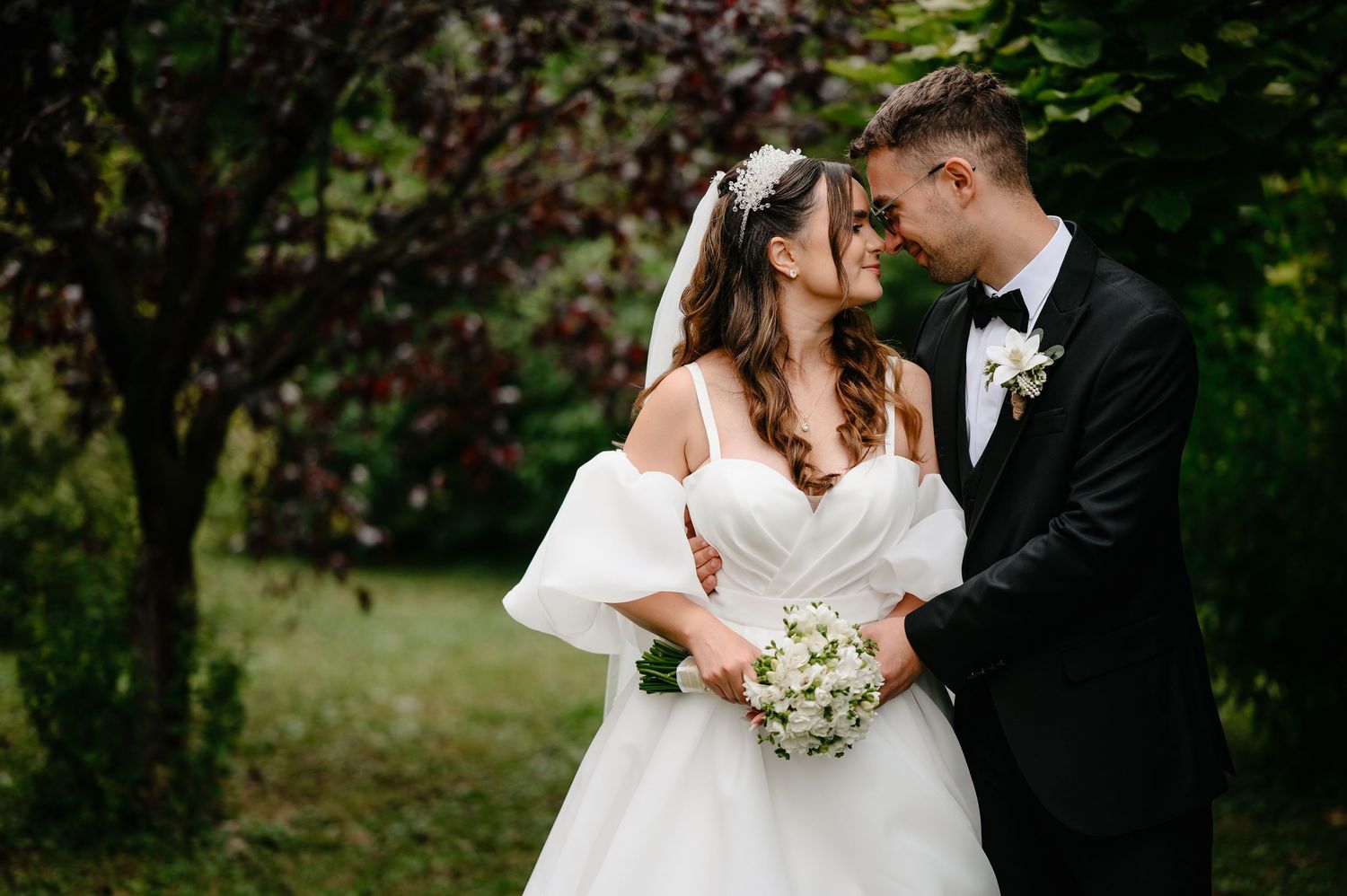 Bride and groom share a romantic forehead touch outdoors surrounded by lush green foliage.