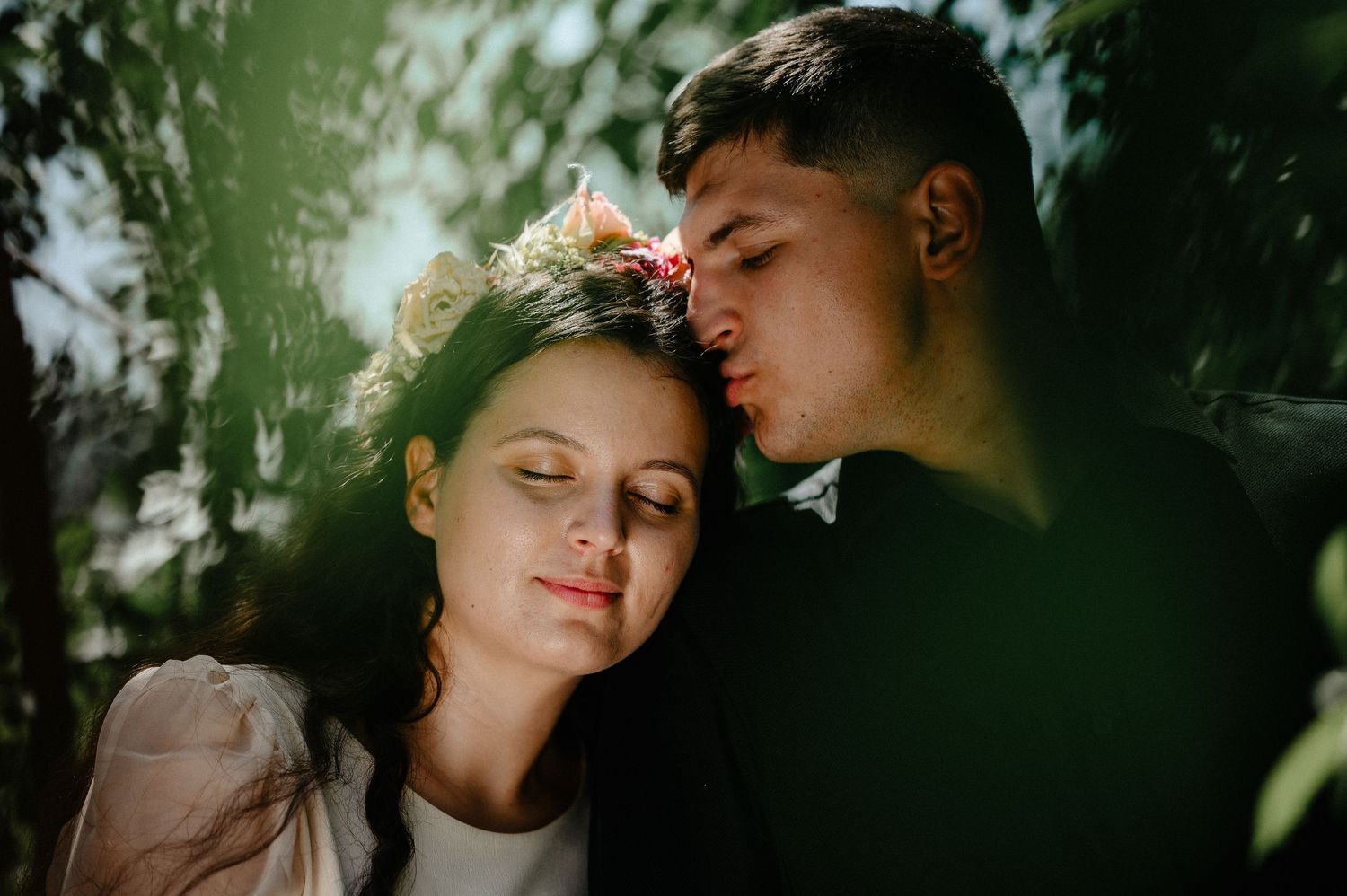 Romantic couple embracing outdoors, woman with flower crown resting head on man's shoulder in sunlit garden.