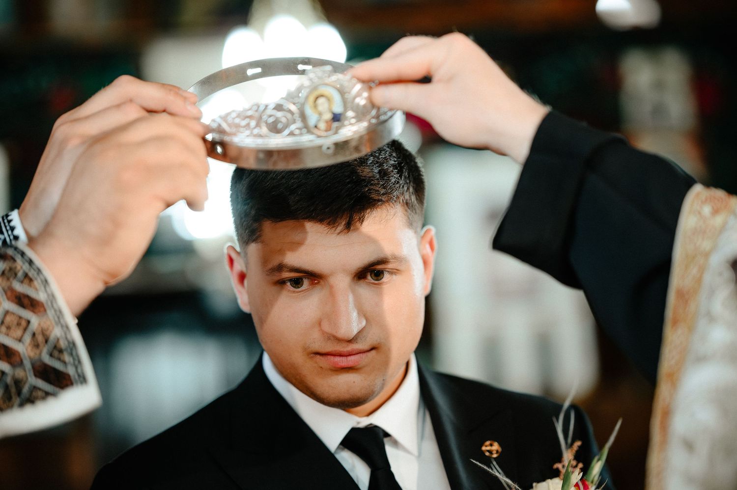 Groom in black suit receiving orthodox wedding crown ceremony from priest.