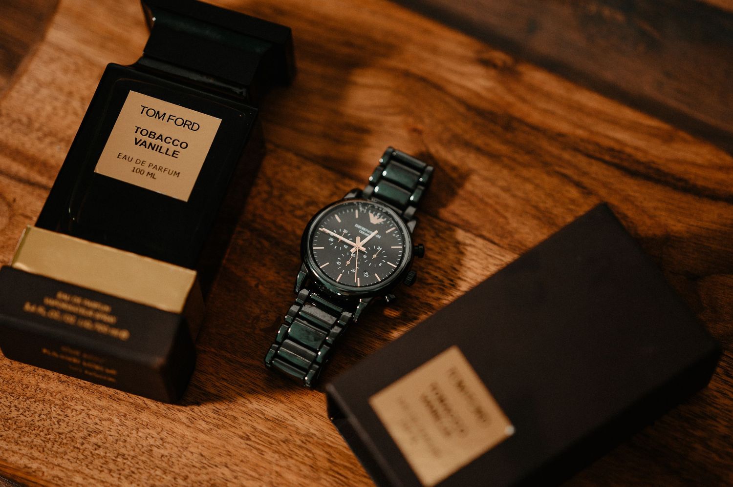 Black chronograph watch beside Tom Ford Tobacco Vanille perfume on wooden surface.