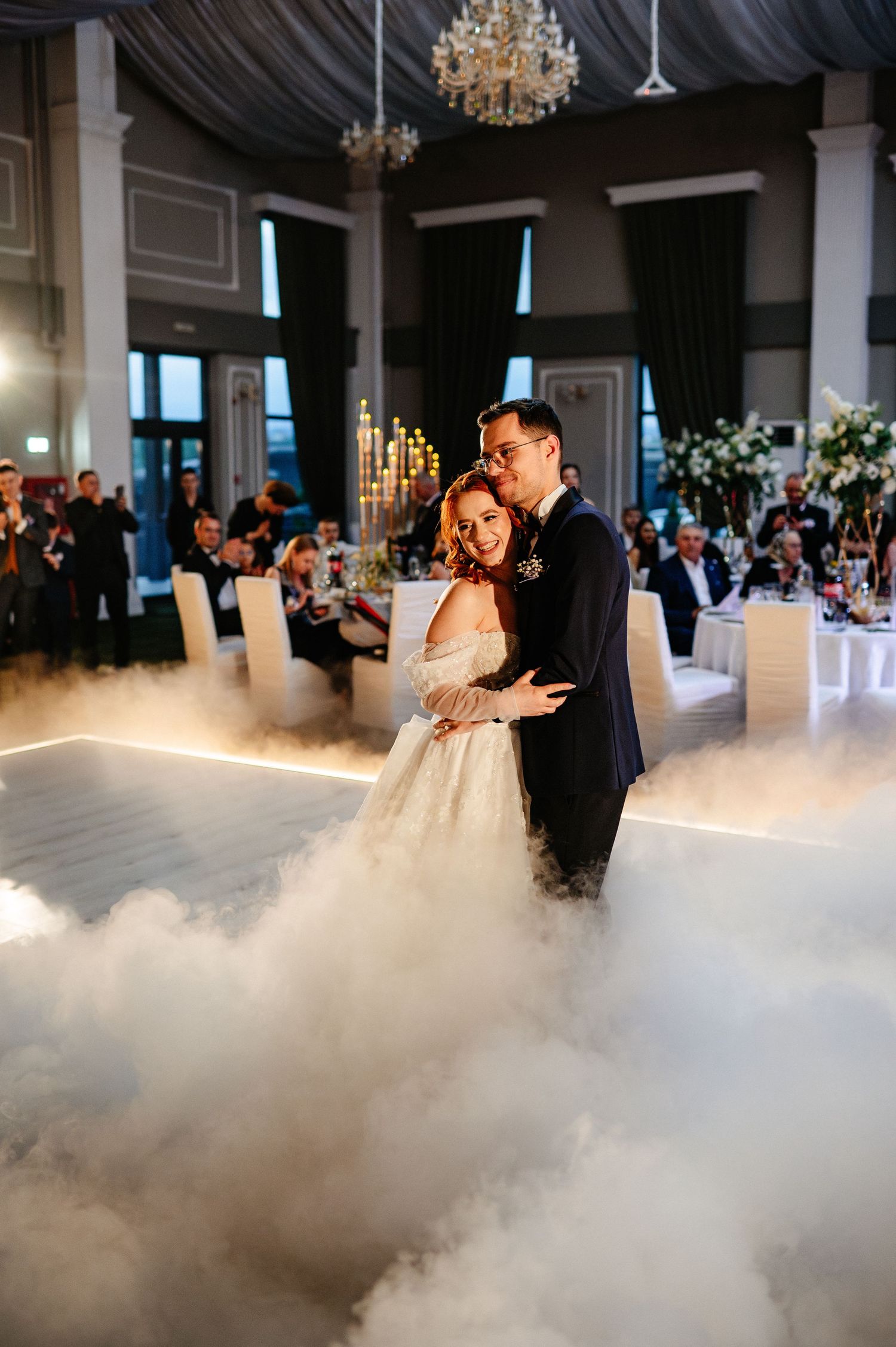 Bride and groom share first dance on fog-filled dance floor in elegant wedding reception venue with chandeliers.