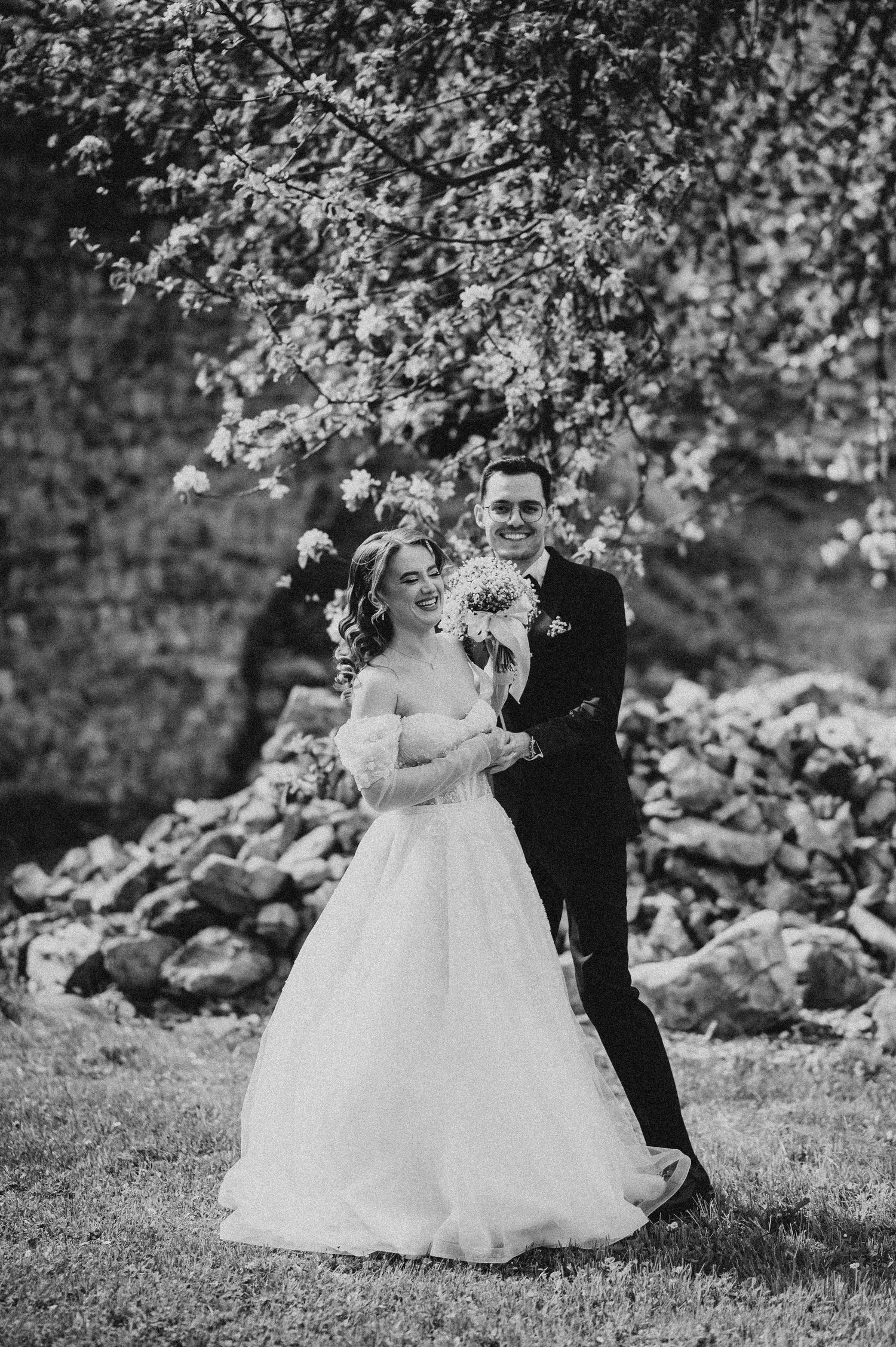 Black and white wedding photo of smiling bride and groom standing under blooming tree with stone wall backdrop.