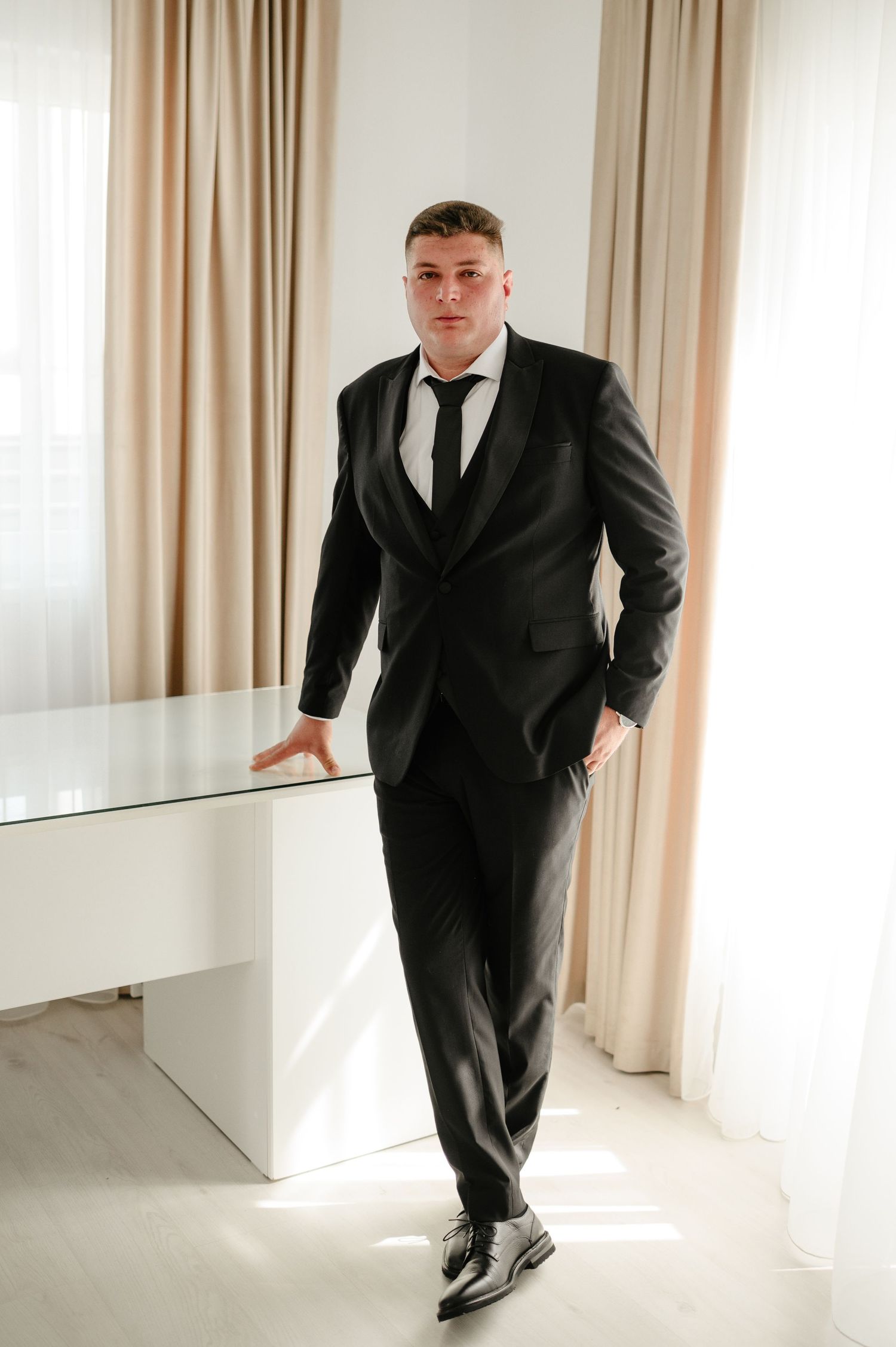 Groom in black suit and tie leaning against white desk in bright room.