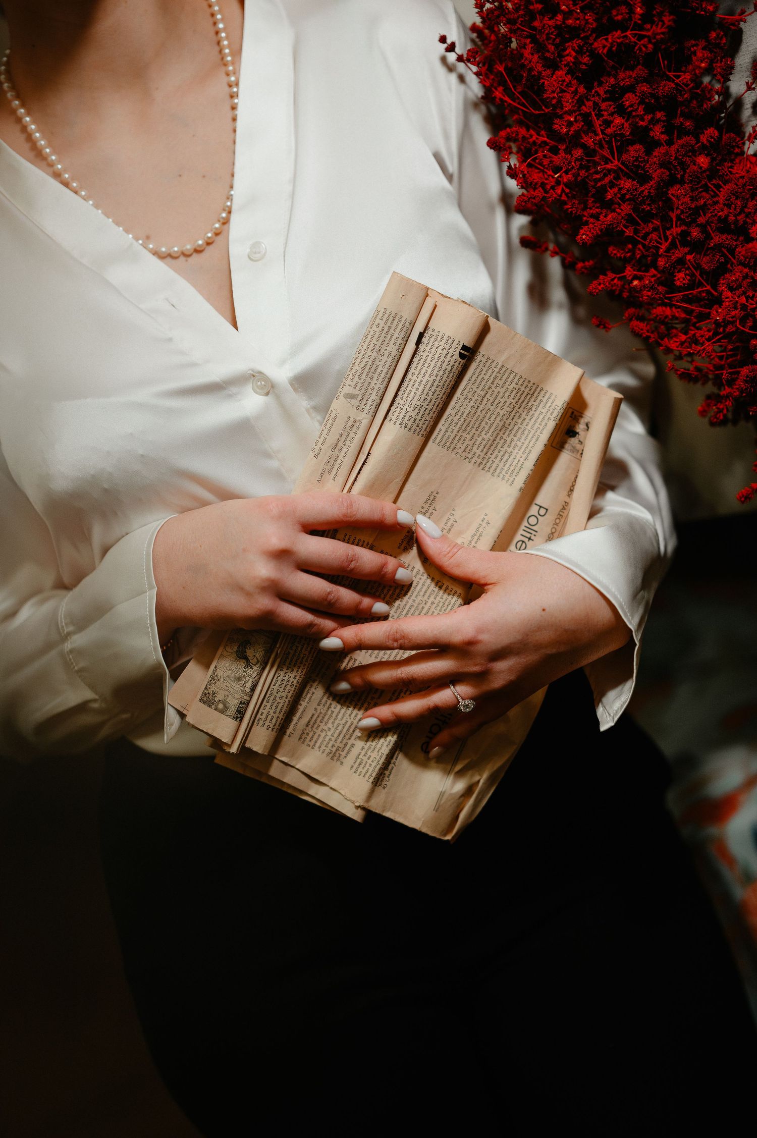 Woman in white blouse holding aged vintage books with red flowers in background, wearing pearl necklace.