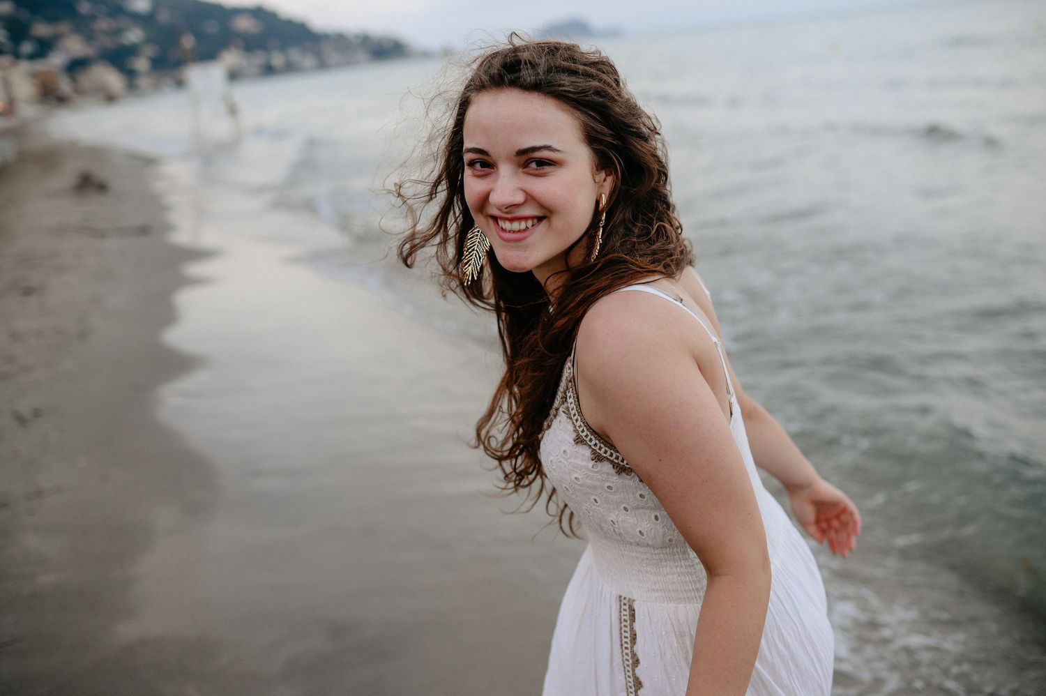 Smiling young woman in white dress posing on a beach with ocean backdrop.