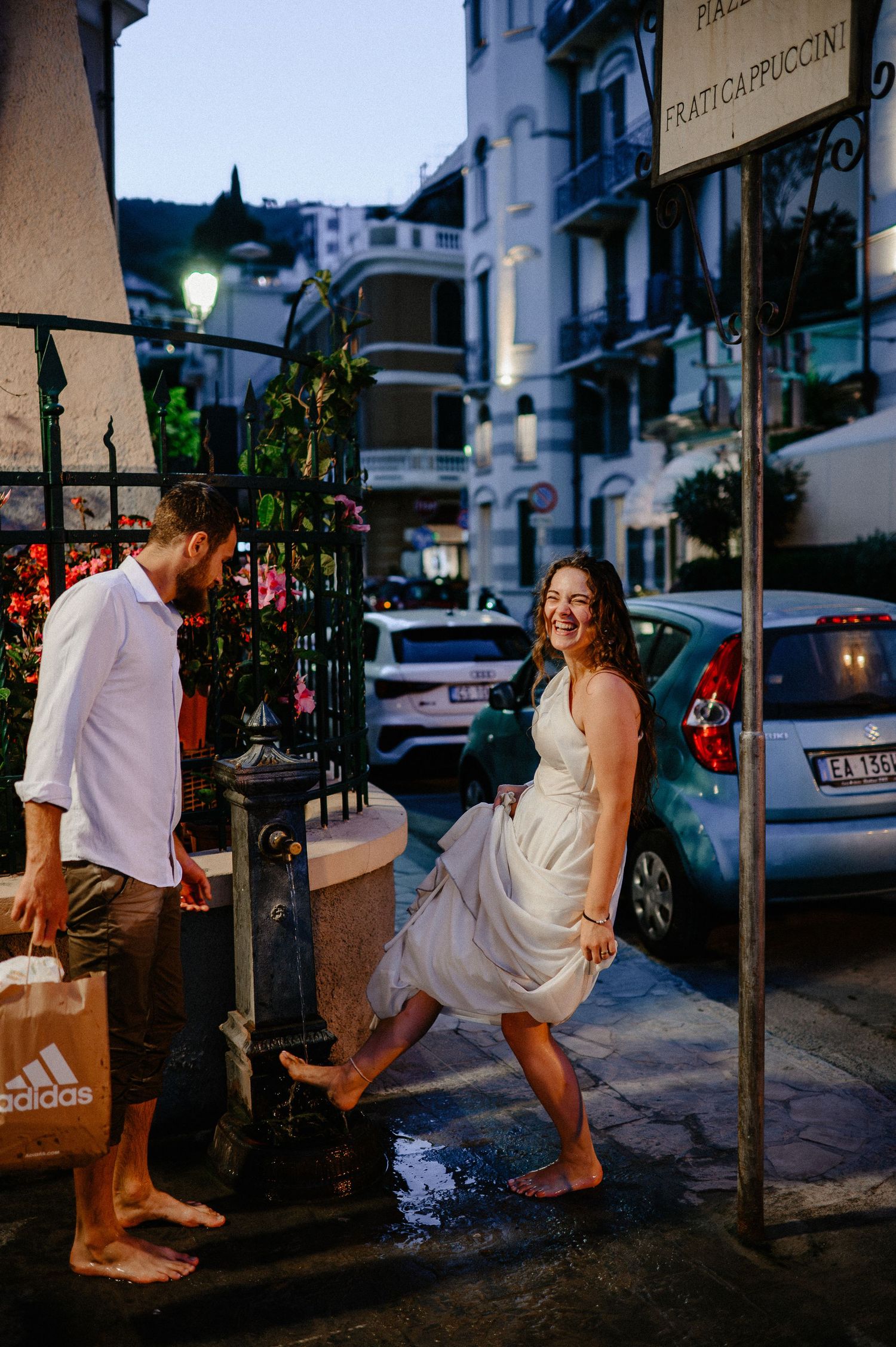 Smiling barefoot woman in white dress stepping through a puddle on a charming Italian street at dusk.