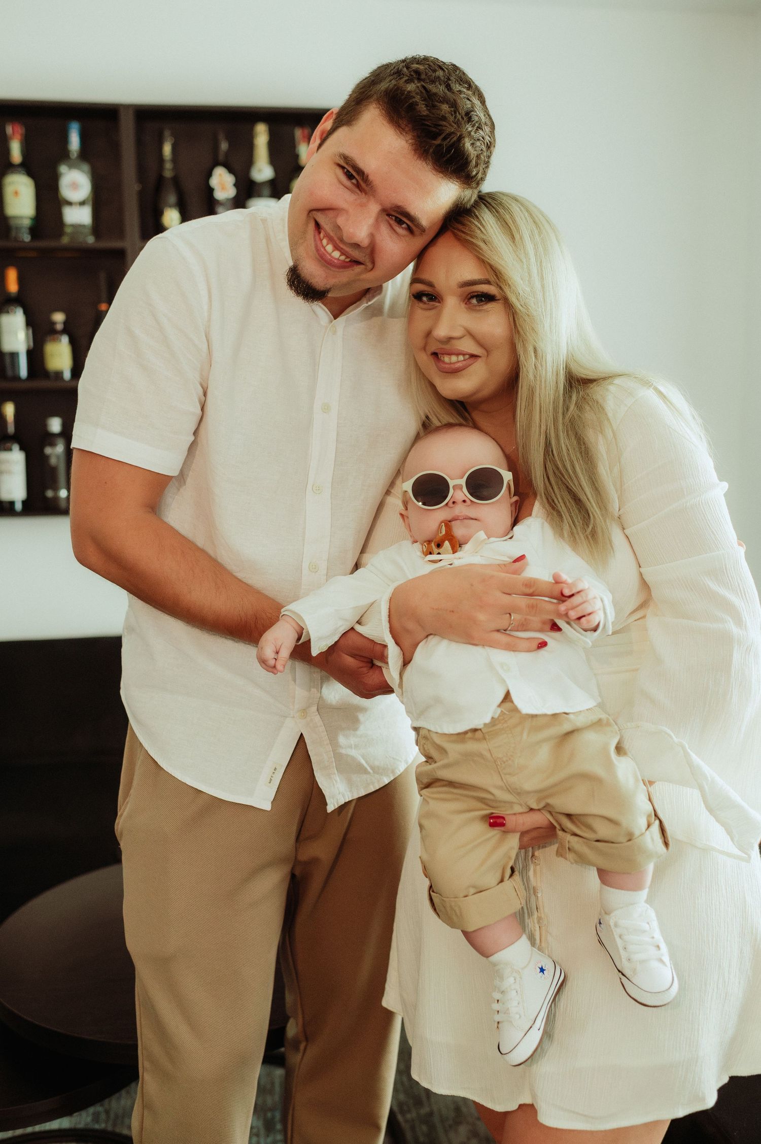 Happy young family portrait with blonde mother holding baby in sunglasses and father smiling.