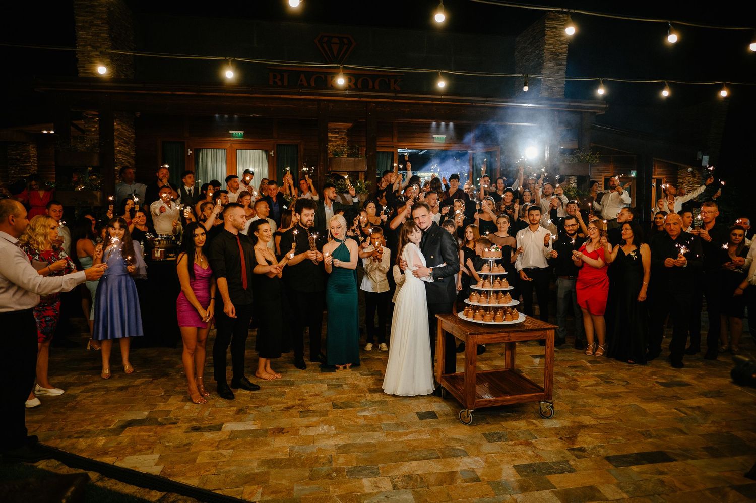 Wedding couple cuts cake surrounded by cheering guests holding sparklers at outdoor reception.