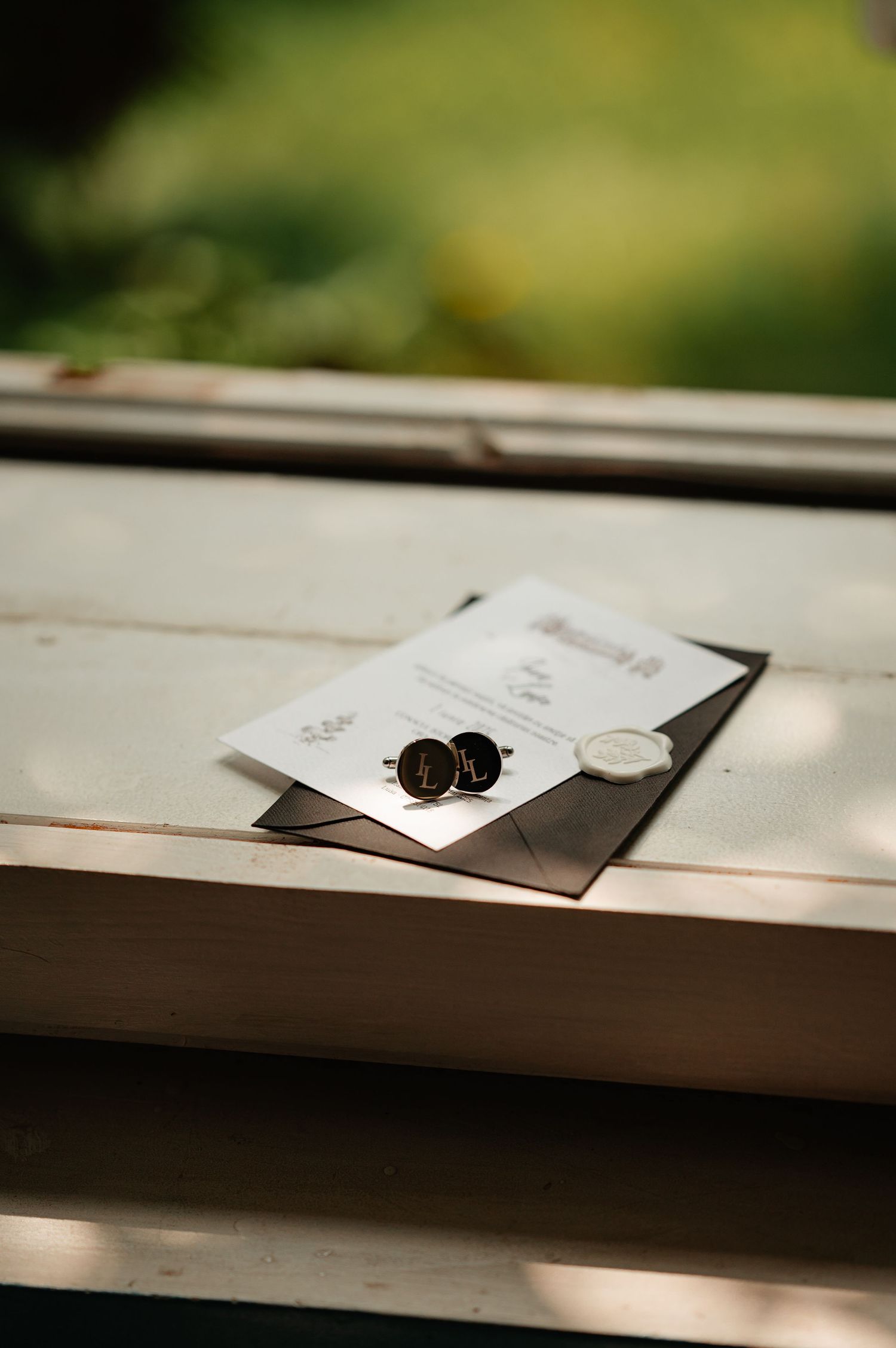 Groom wedding cufflinks resting on elegant invitation with wax seal on wooden window ledge.