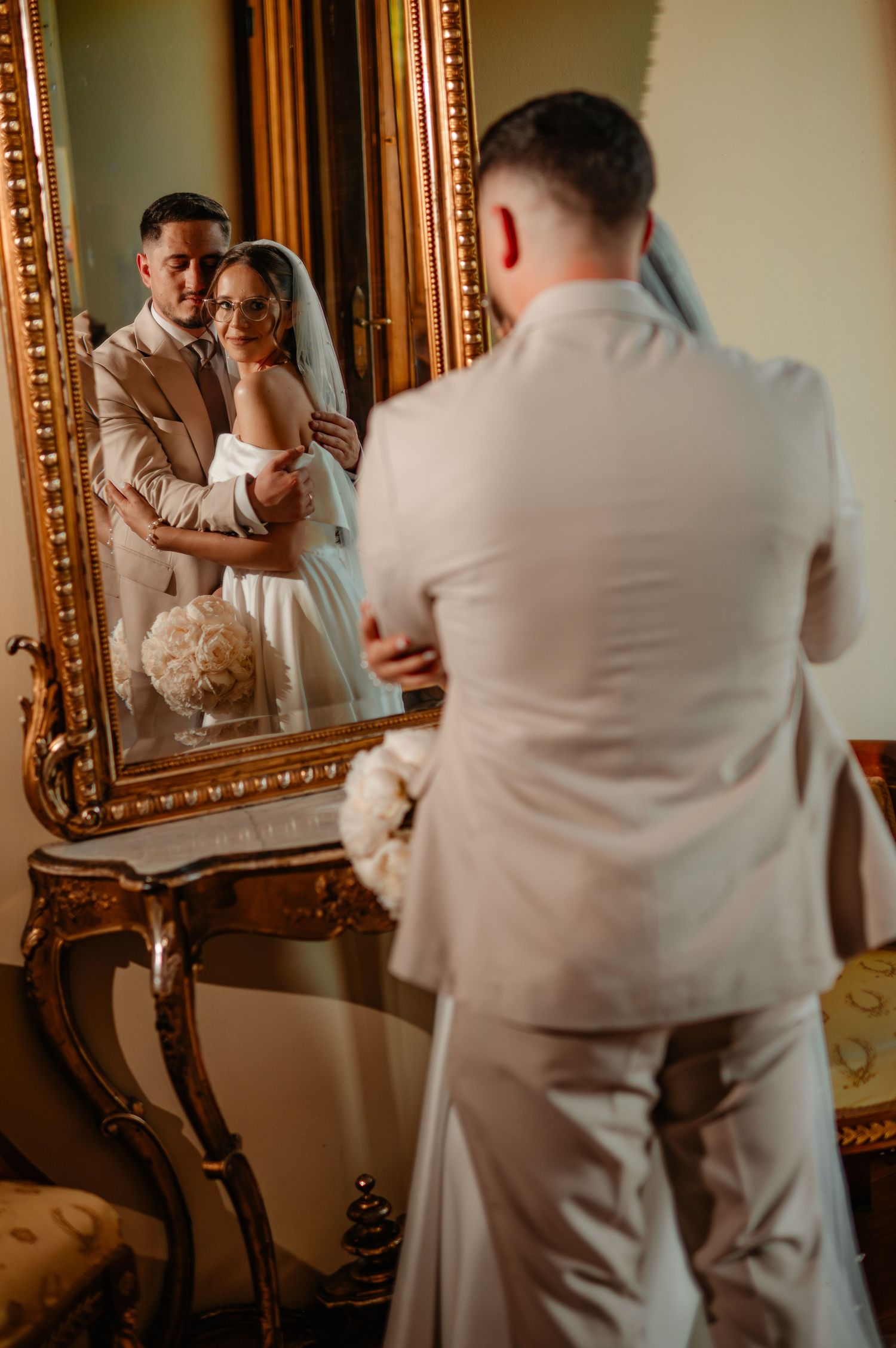 Groom embraces bride from behind as their romantic reflection is captured in an ornate gold-framed vintage mirror.