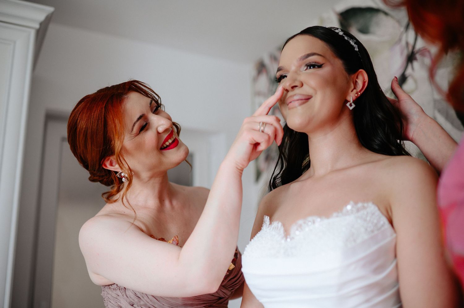 Bridesmaid applying makeup to smiling bride in white strapless wedding dress during bridal preparations.