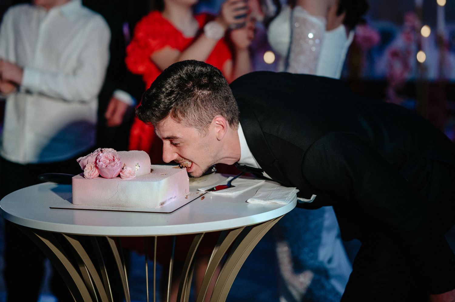 Groom in black suit leans down to take a bite directly from the pink wedding cake on table.