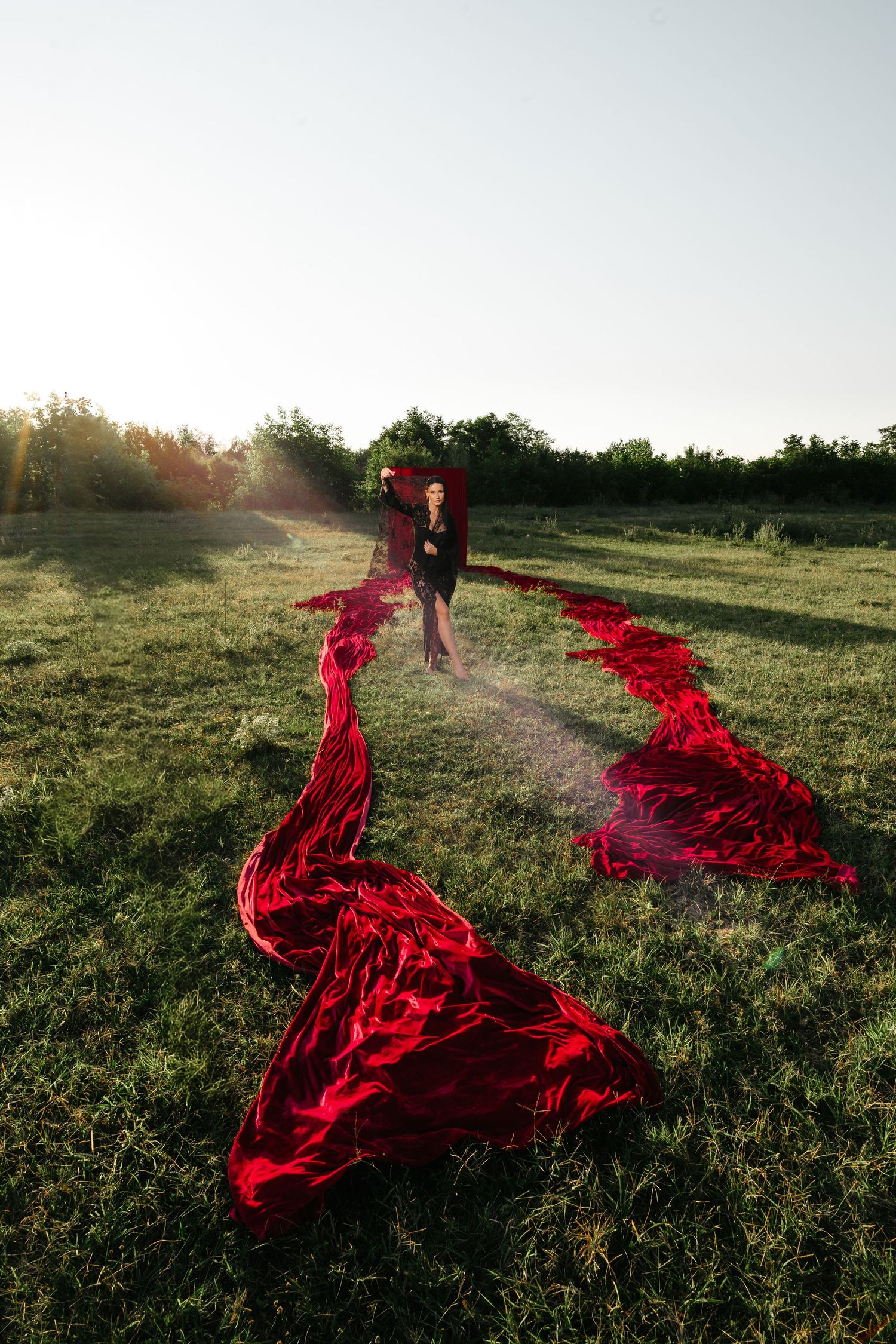 Woman in black dress holding flowing red fabric in sunlit green field.