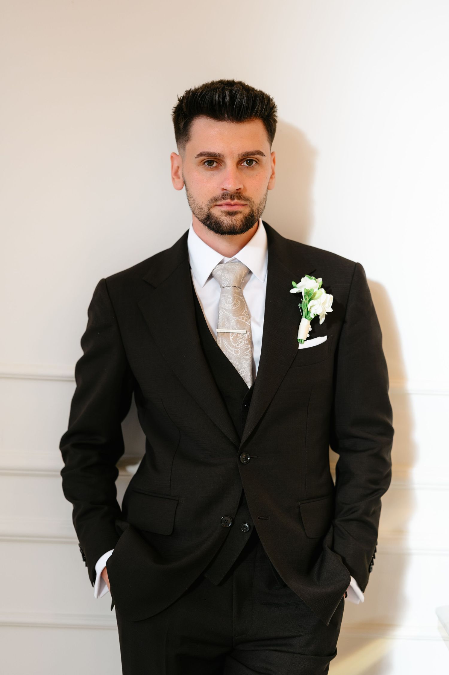 Groom in black three-piece suit with floral boutonniere and beige tie poses confidently against white wall.