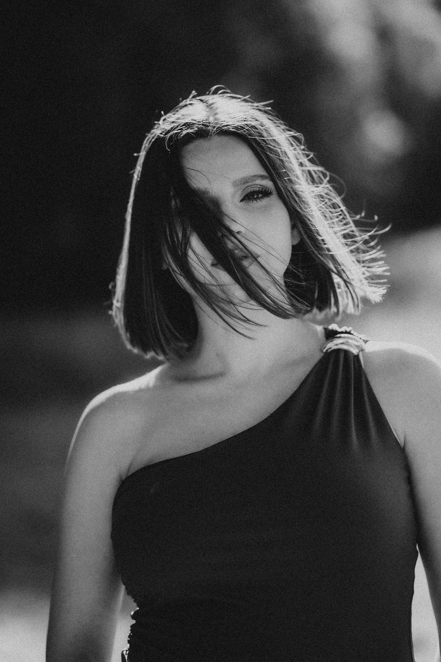 Black and white portrait of woman in one-shoulder dress with windswept hair outdoors.