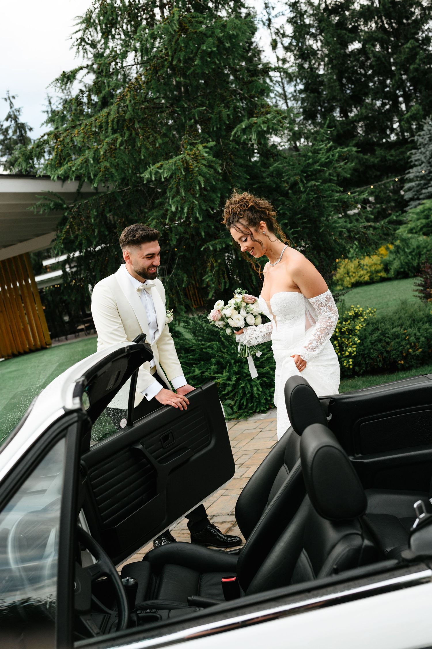 Bride and groom in white attire stand beside a convertible car surrounded by lush greenery on their wedding day.