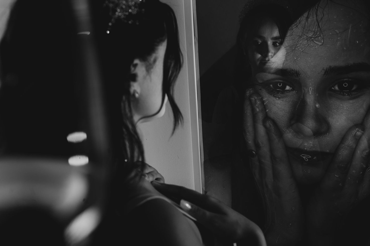 Black and white photo of a woman touching her face reflected in a rain-covered mirror.