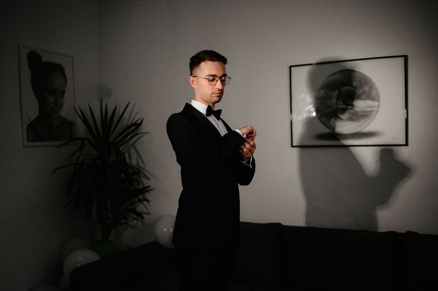 A man in a black tuxedo with bow tie adjusts his cufflinks in a dimly lit room with framed art.