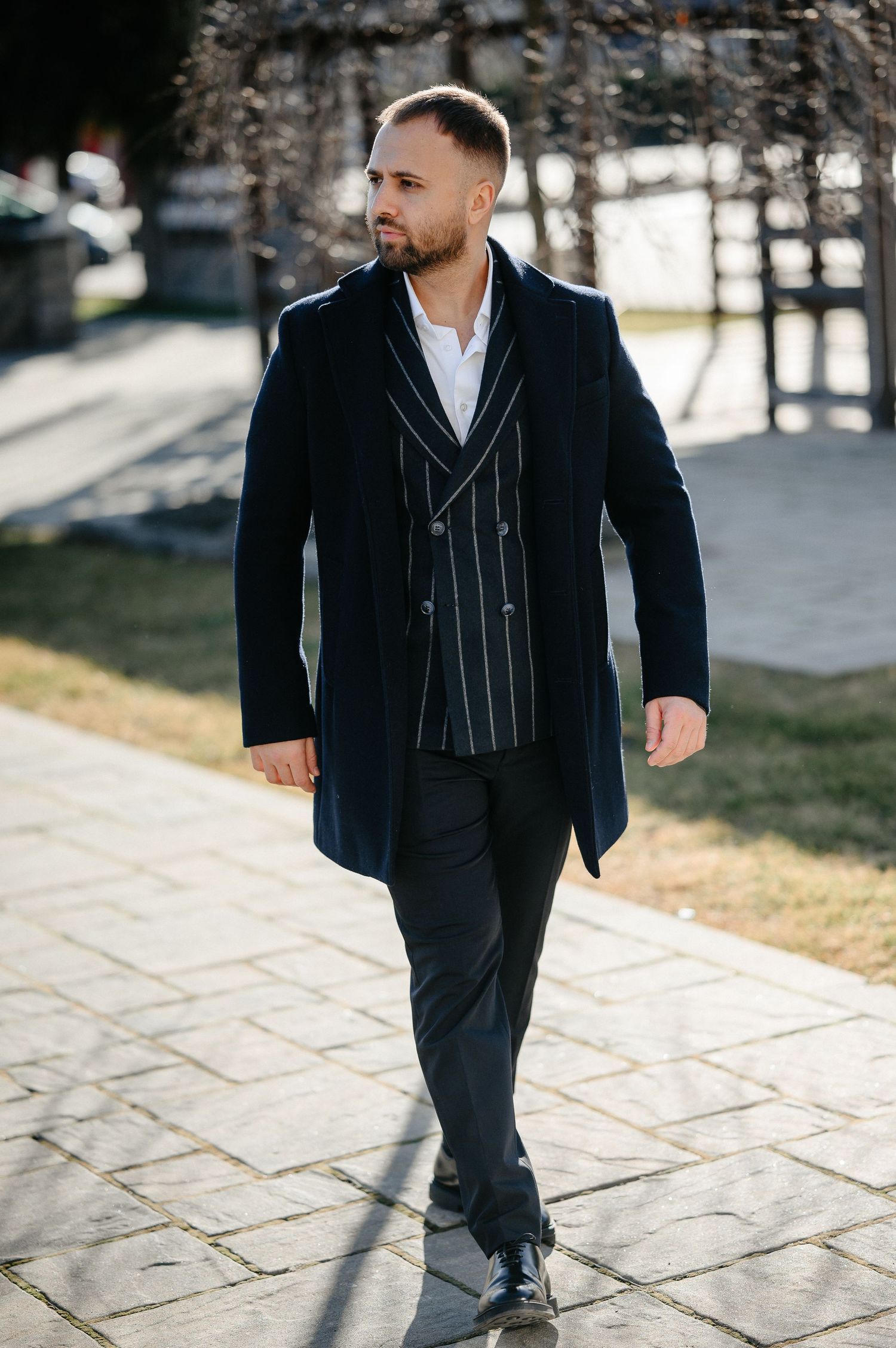 Stylish groom in navy double-breasted coat and striped vest walks confidently on stone path.