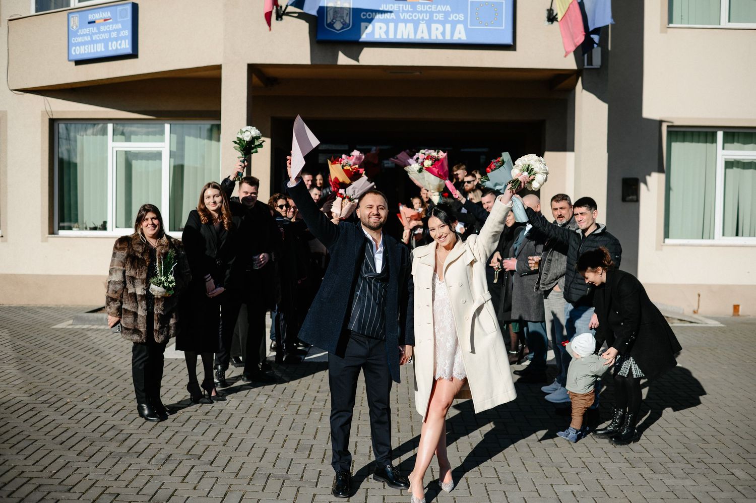 Joyful wedding party celebrating outside a municipal building with flowers and raised hands.