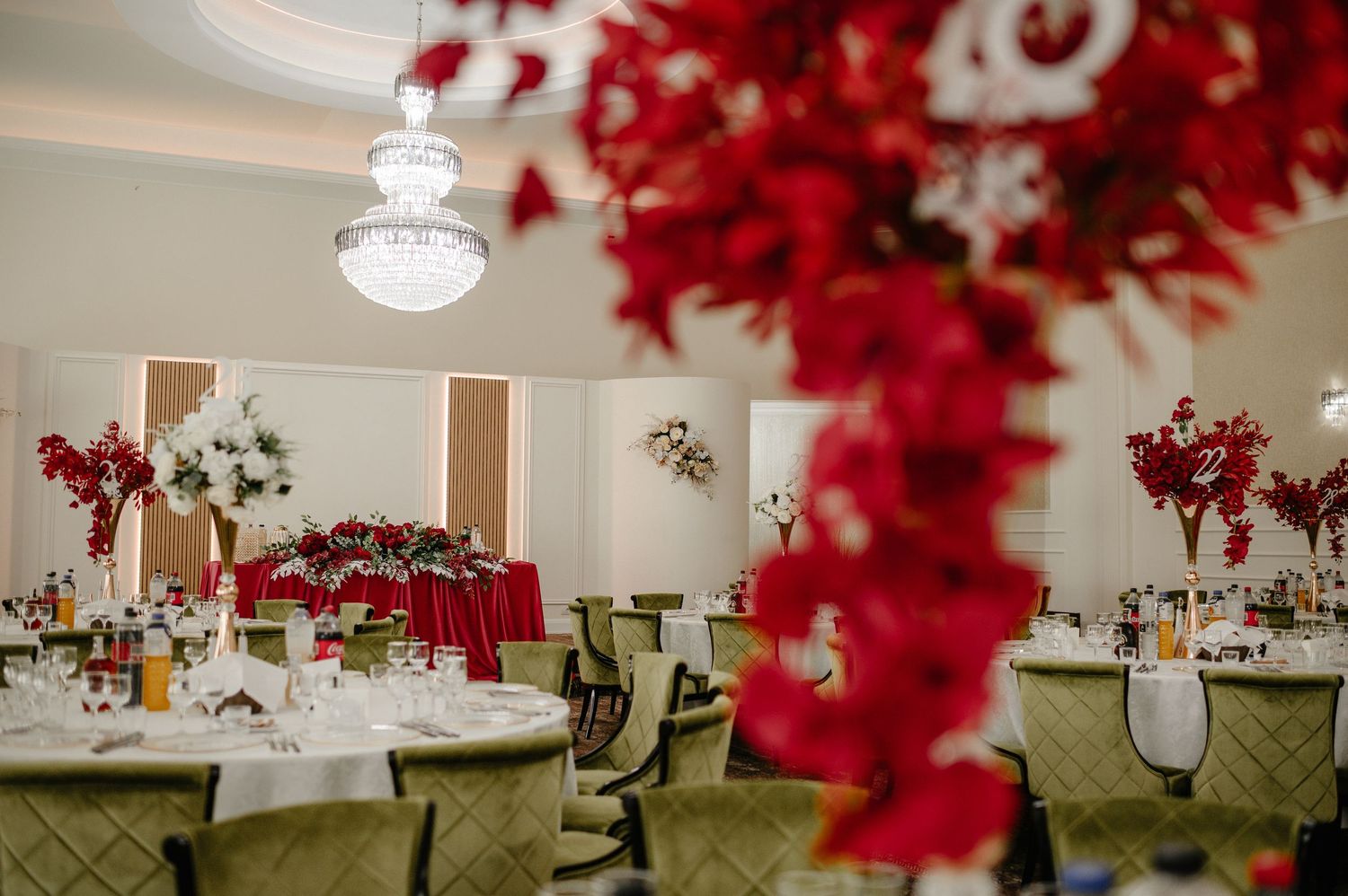 Elegant wedding reception hall decorated with tall red floral centerpieces and round tables with green chairs.