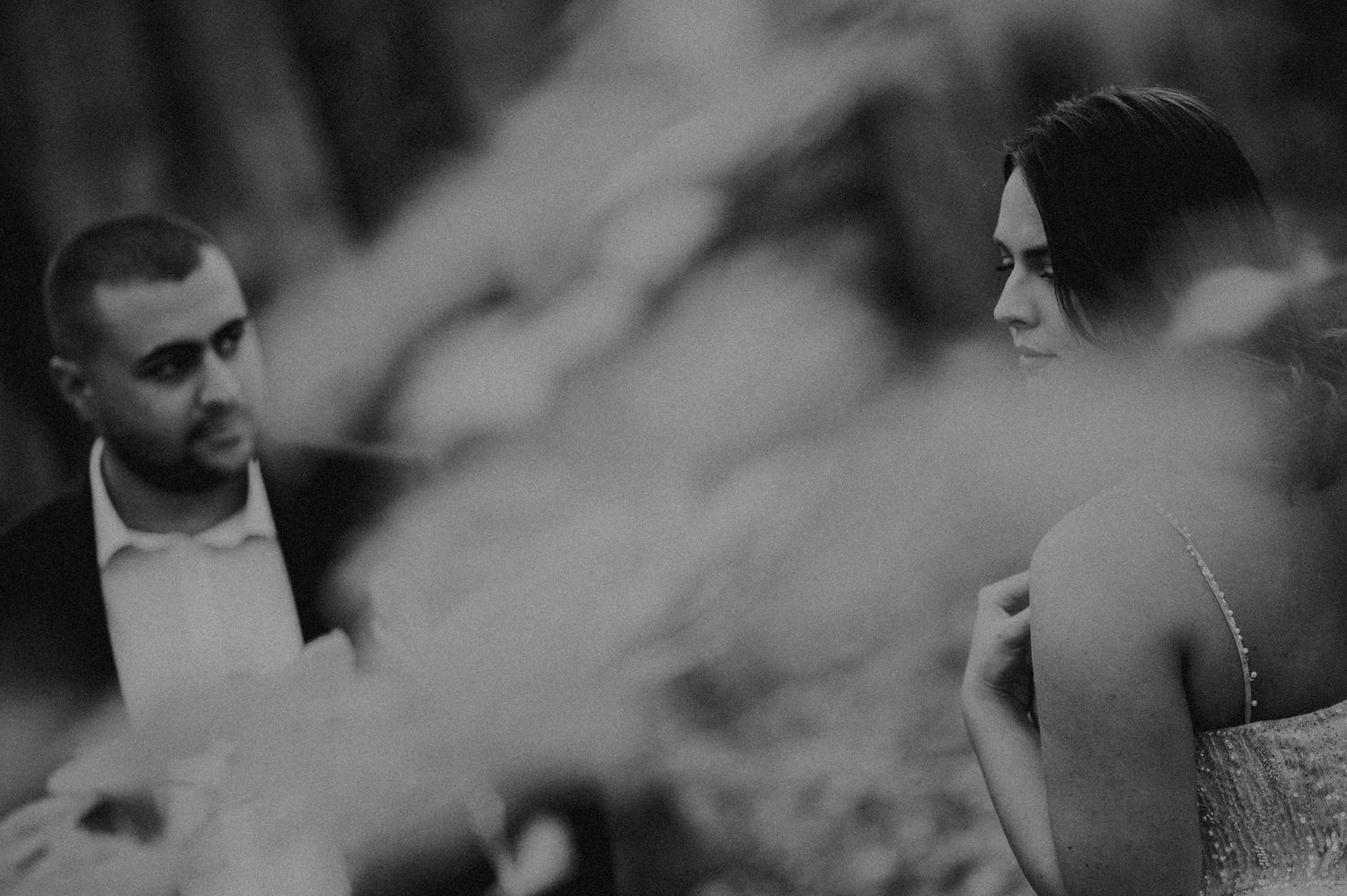 Black and white wedding photo of bride and groom standing outdoors, with soft bokeh foliage in foreground.