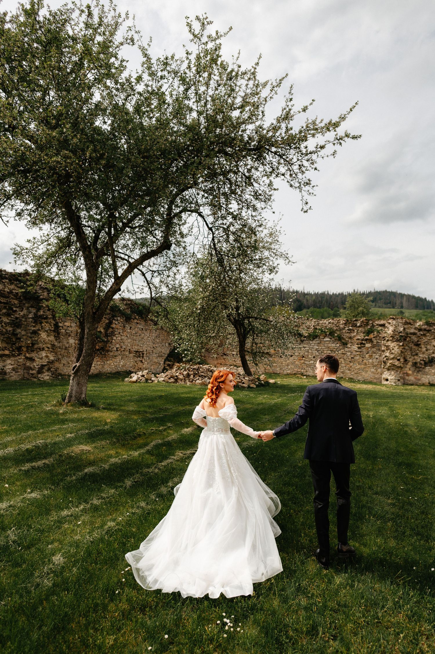 A bride in a white gown and groom in a dark suit hold hands walking through a lush green garden near stone ruins.