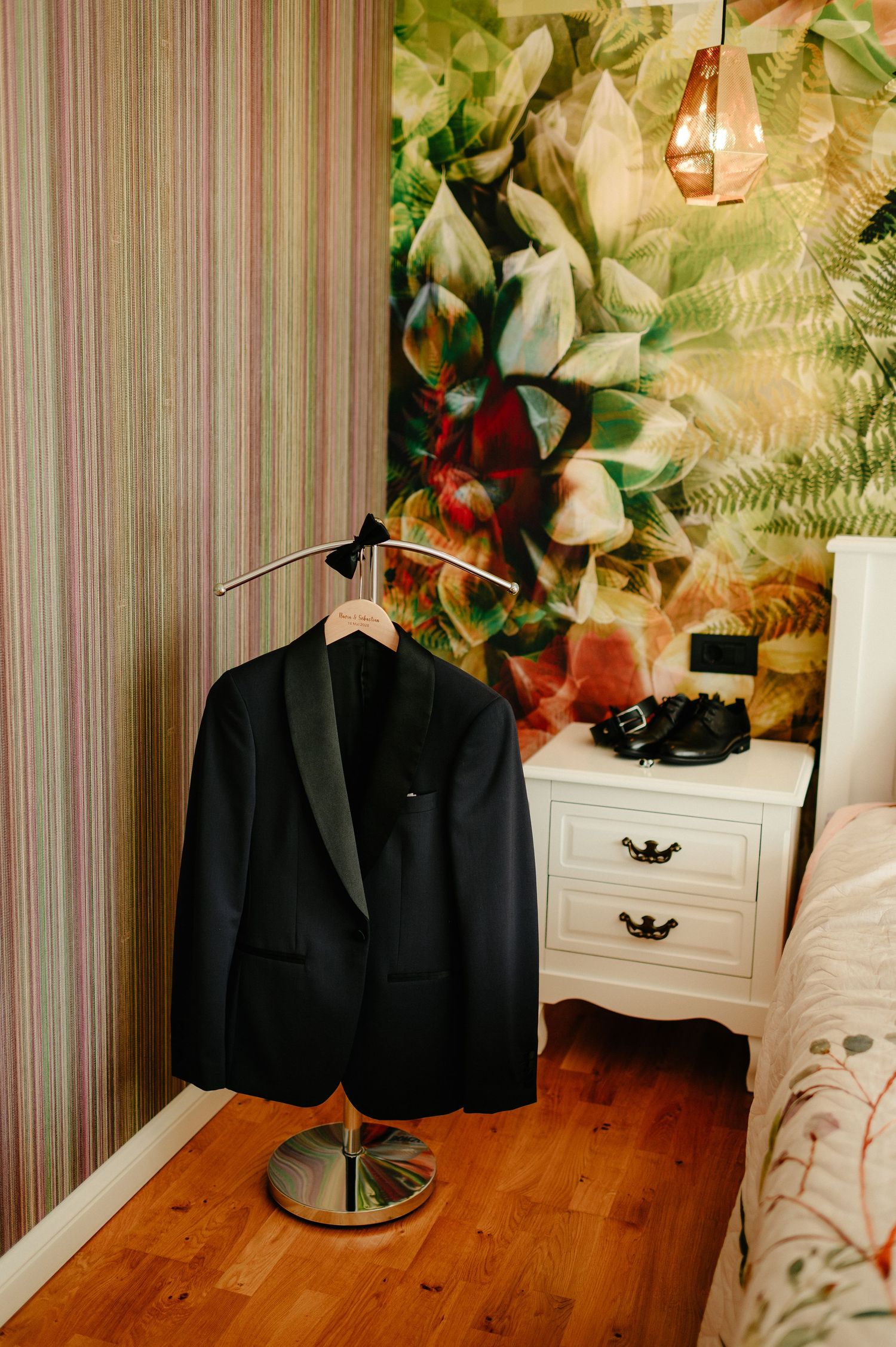 Black tuxedo jacket on a freestanding hanger in a stylish bedroom with tropical wallpaper and white nightstand.