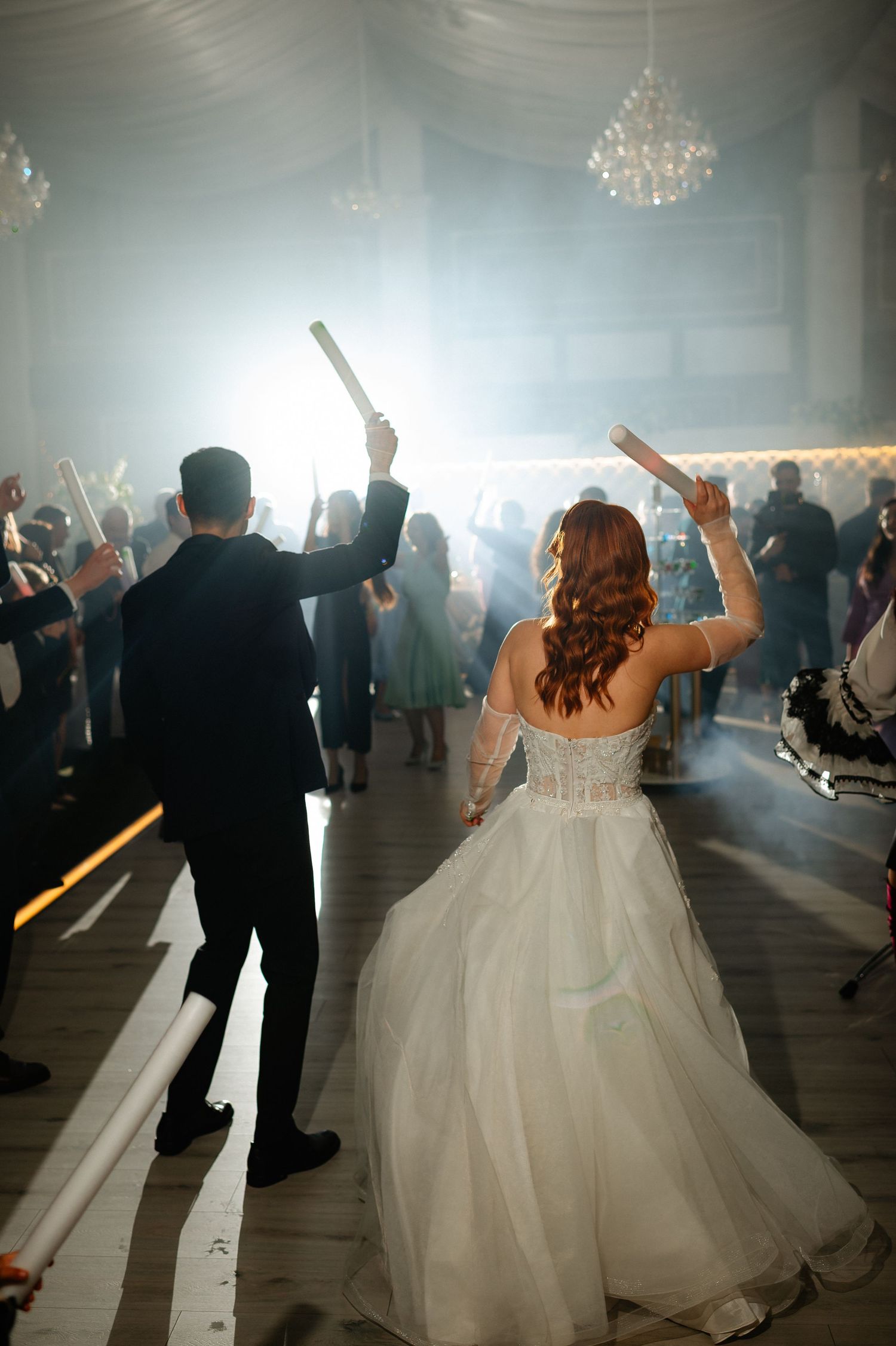 Bride and groom dancing at wedding reception holding glow sticks surrounded by guests in elegant venue with chandeliers.