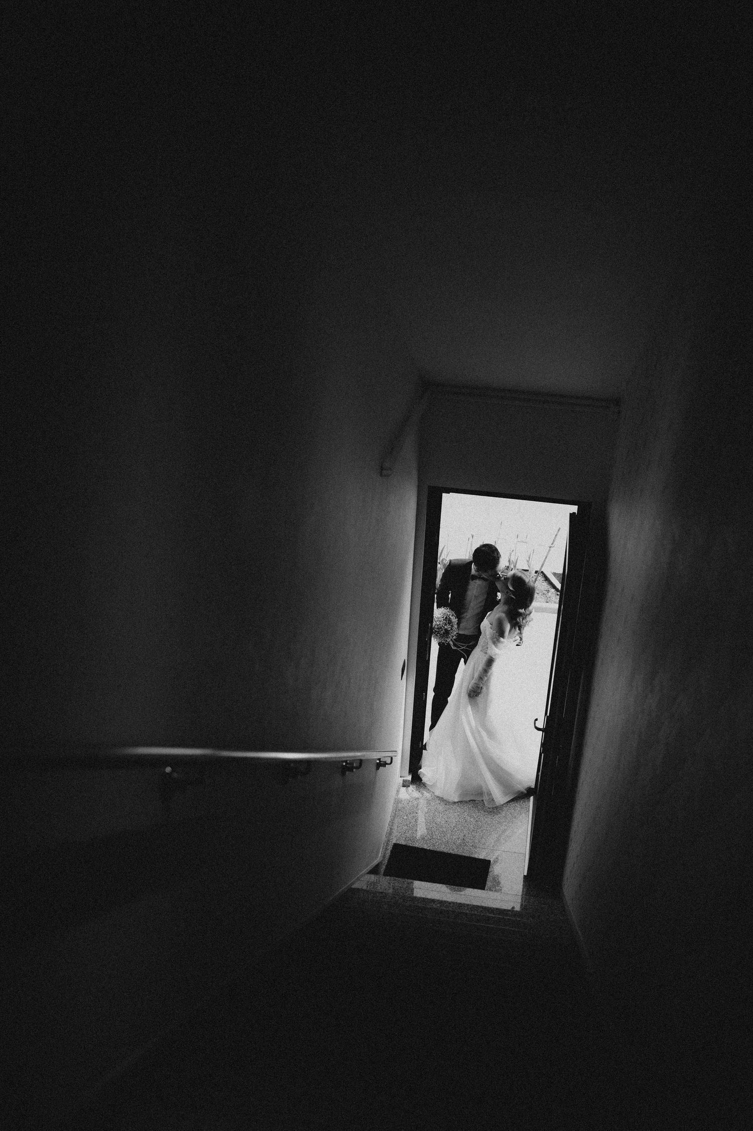 Black and white wedding photo of bride and groom kissing in a doorway, viewed through a dark stairwell.