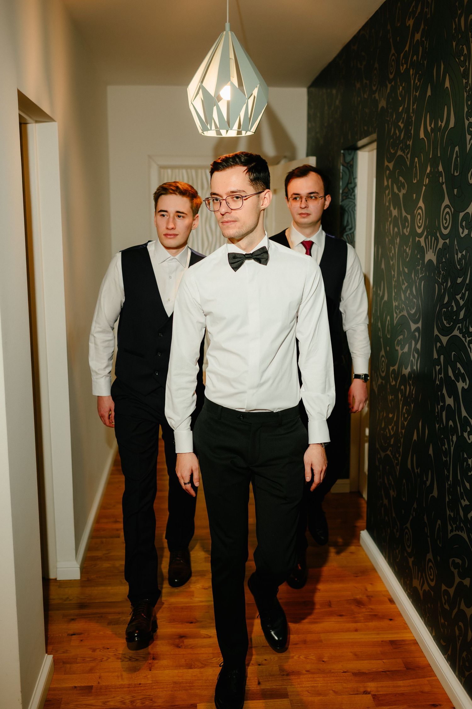 Three well-dressed men in white shirts and black vests walk confidently down a hallway toward the camera.