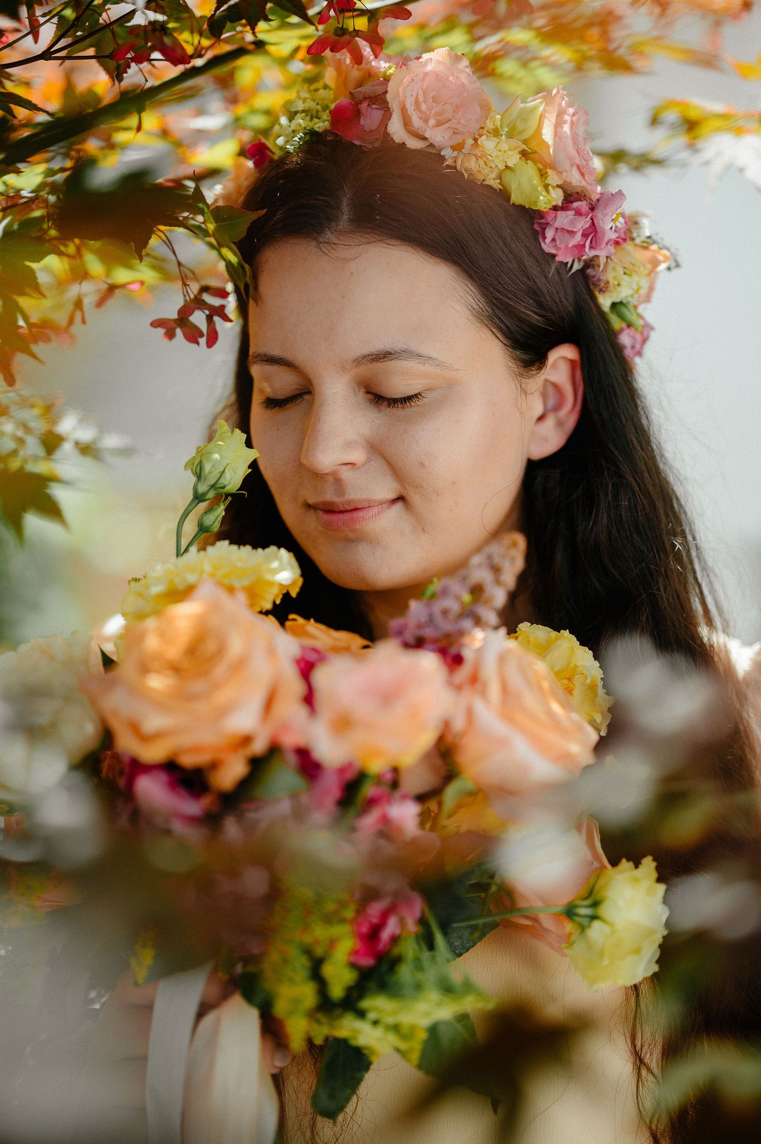 Young woman with floral crown and peach roses bouquet surrounded by autumn foliage in soft natural light.