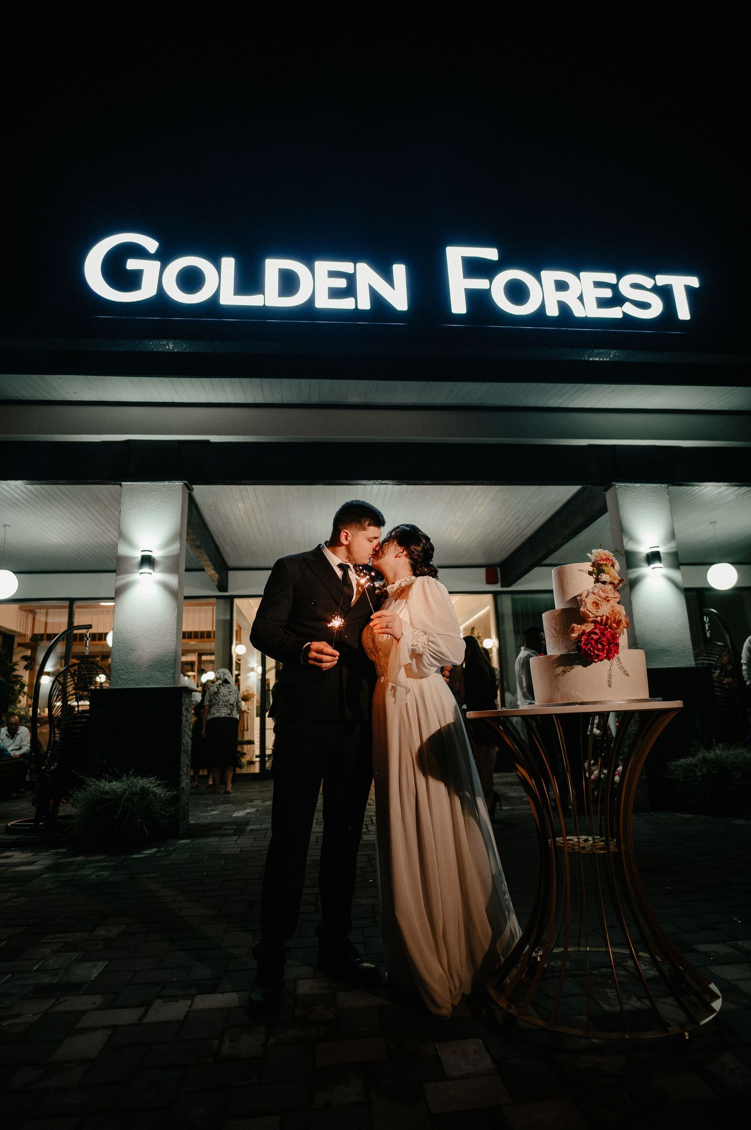 Newlyweds kissing with sparklers outside Golden Forest wedding venue at night.