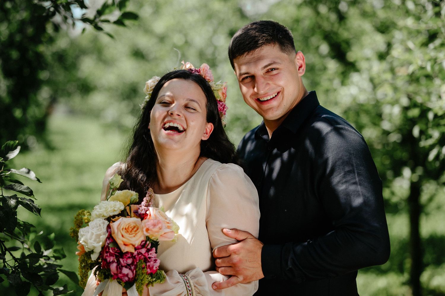 Smiling couple posing outdoors in a green garden, woman holding a colorful bouquet with pink flowers in her hair.