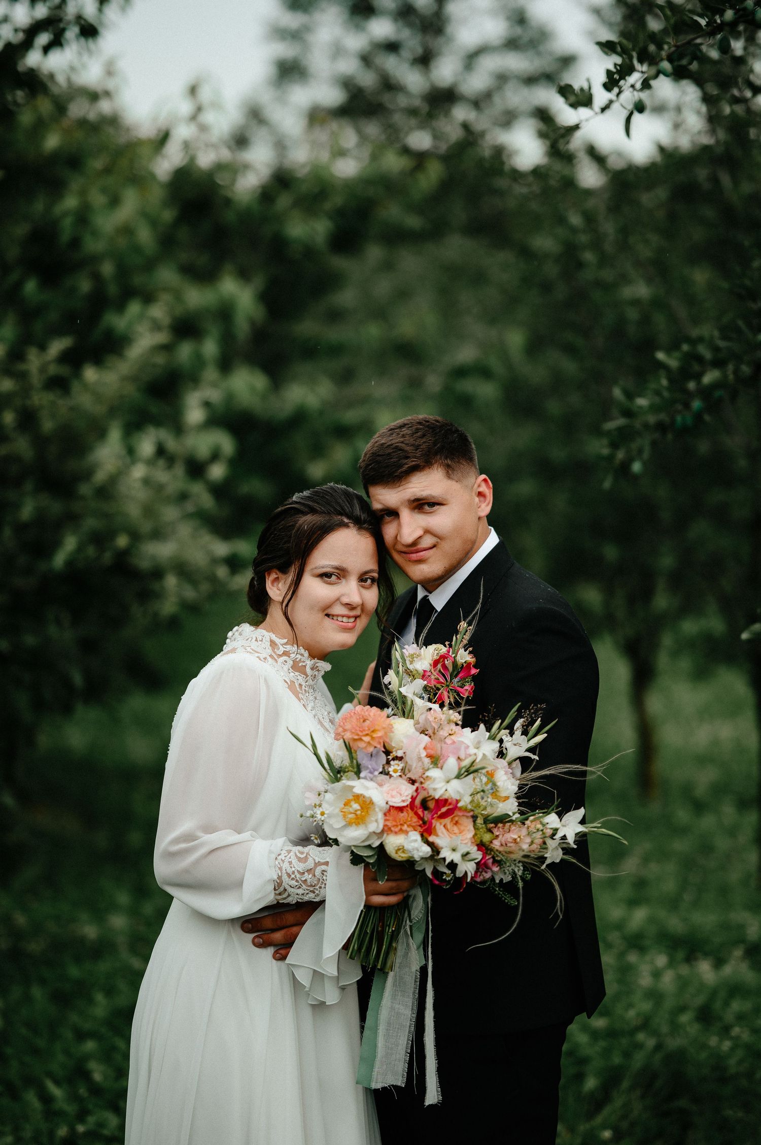 Bride in white lace gown holding colorful bouquet poses with groom in black suit in lush green garden.