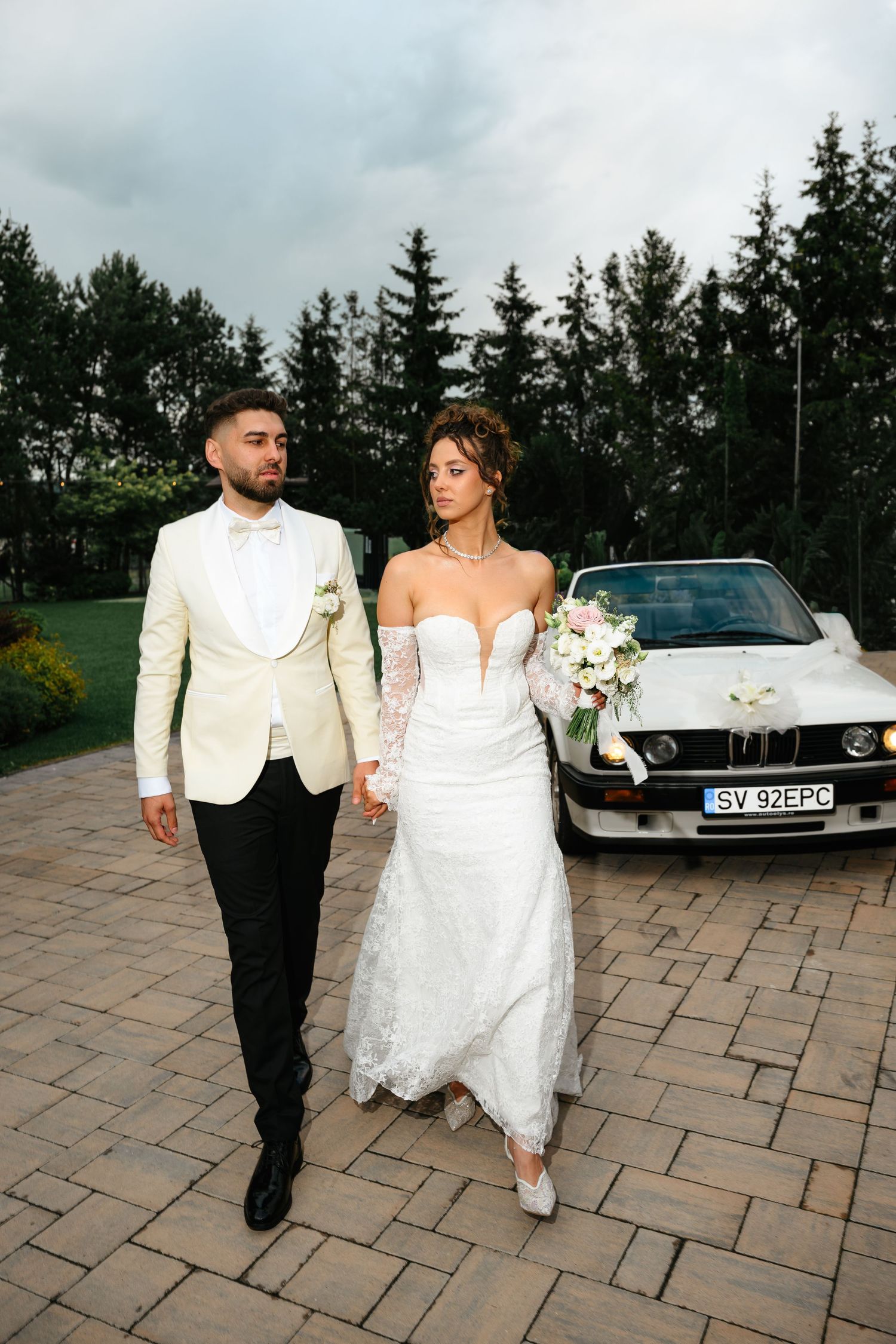 Bride and groom walking hand in hand in front of a white BMW, surrounded by lush green trees on their wedding day.