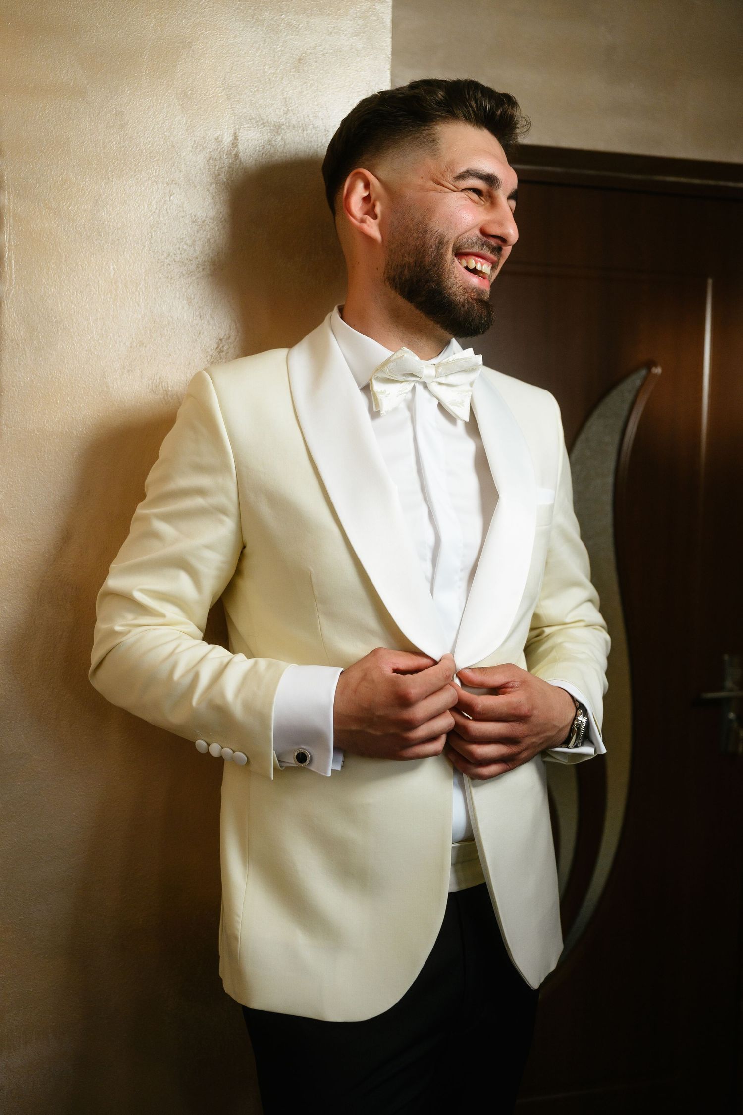 Smiling groom in ivory tuxedo jacket and white bow tie buttoning his jacket while leaning against a wall.