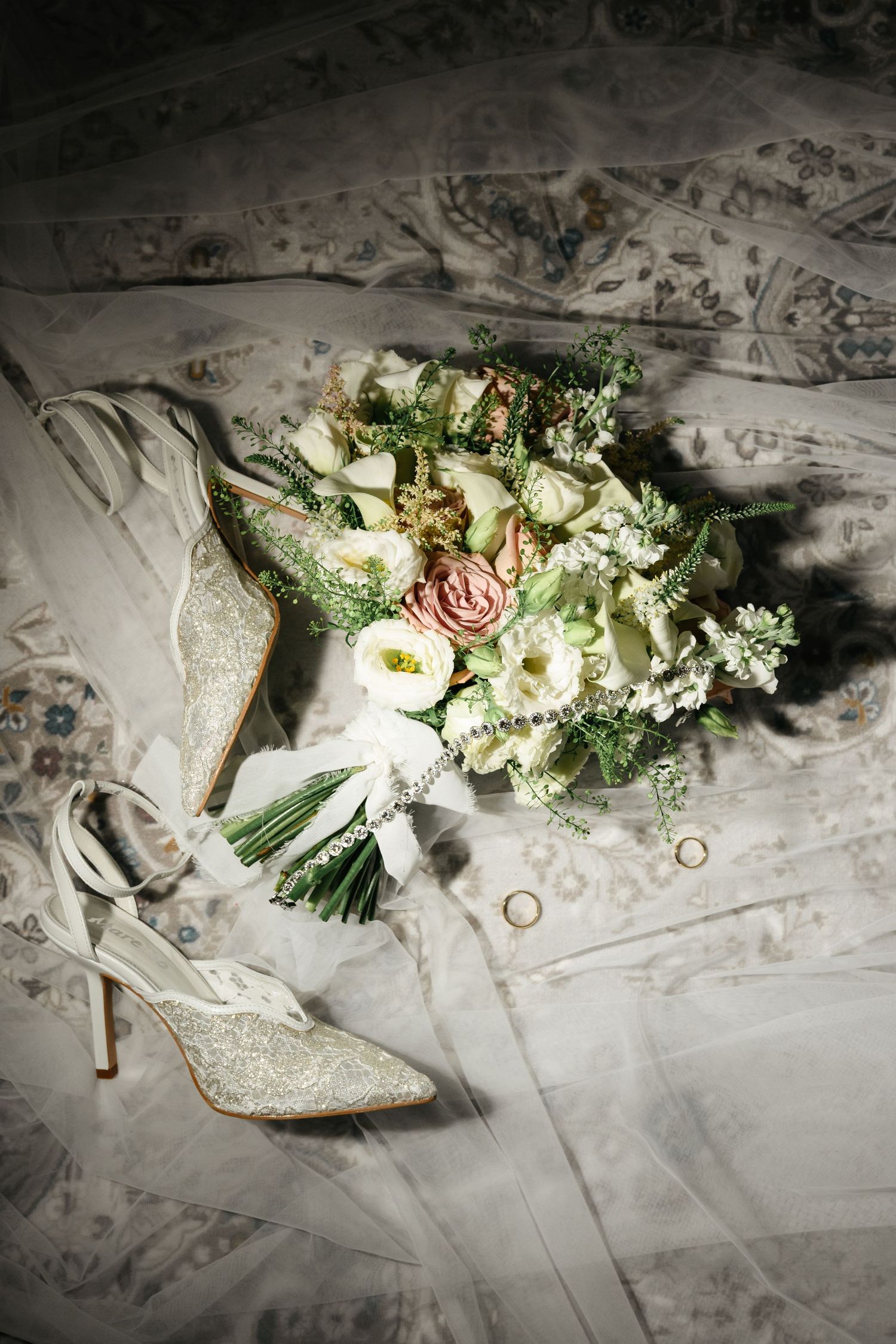 Elegant bridal flat lay with lace heels, wedding bouquet of roses and greenery, and gold rings on white veil.